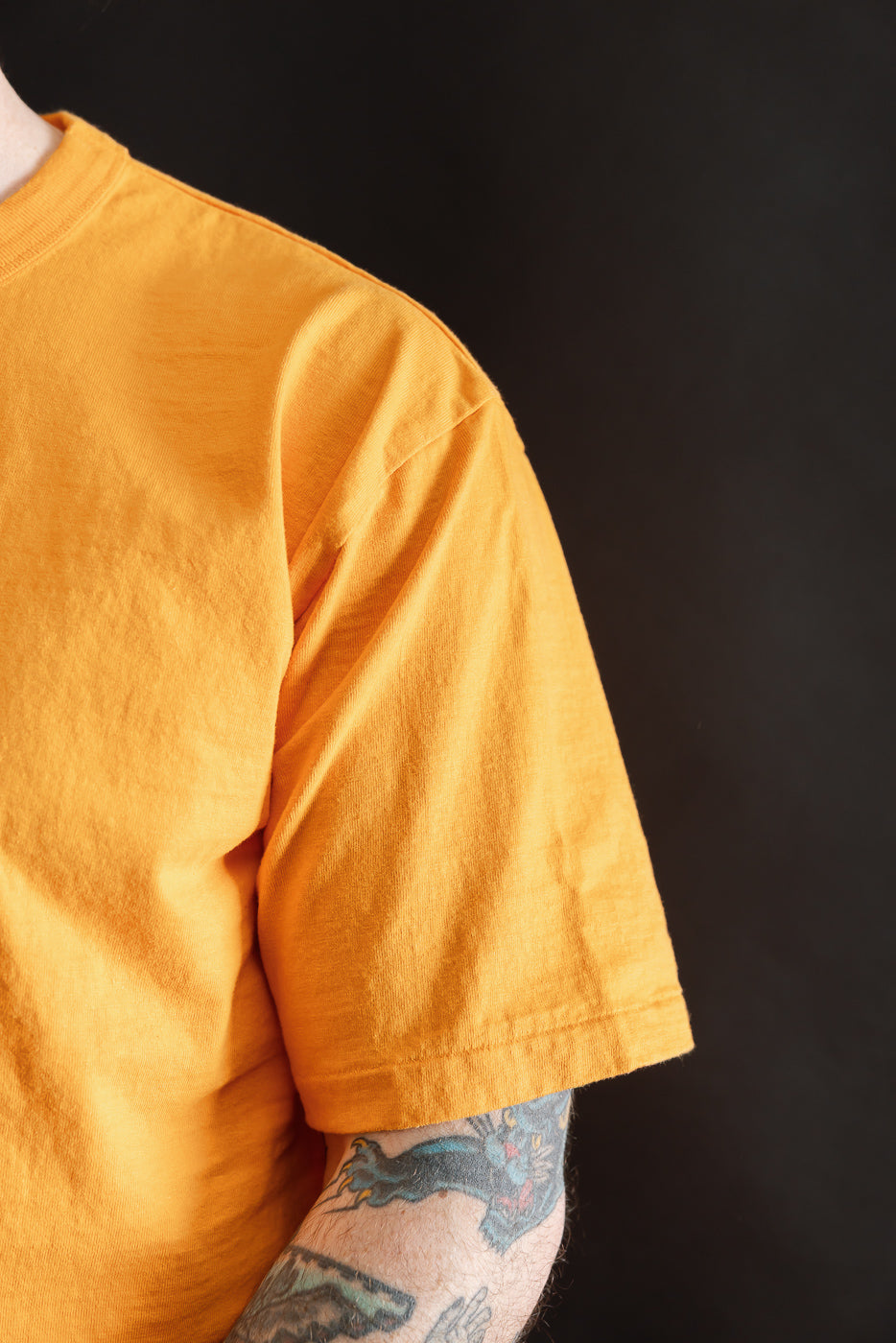 Close-up of the 7.7oz Fallen Cotton Midweight T-Shirt S/S - Fade Orange, highlighting the mustard-yellow fabric and colorful tattoos on the right forearm, set against a dark backdrop.