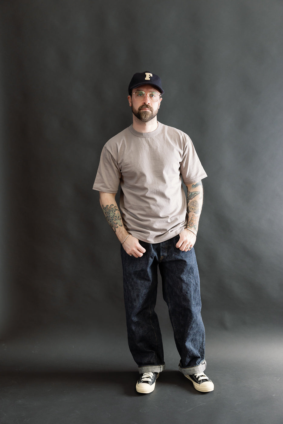 Man in Fallen Cotton Midweight T-Shirt, grey, styled with loose-fit dark denim jeans and low-top sneakers, showcasing a casual look. The model has a relaxed stance, highlighting the tee's comfortable fit.