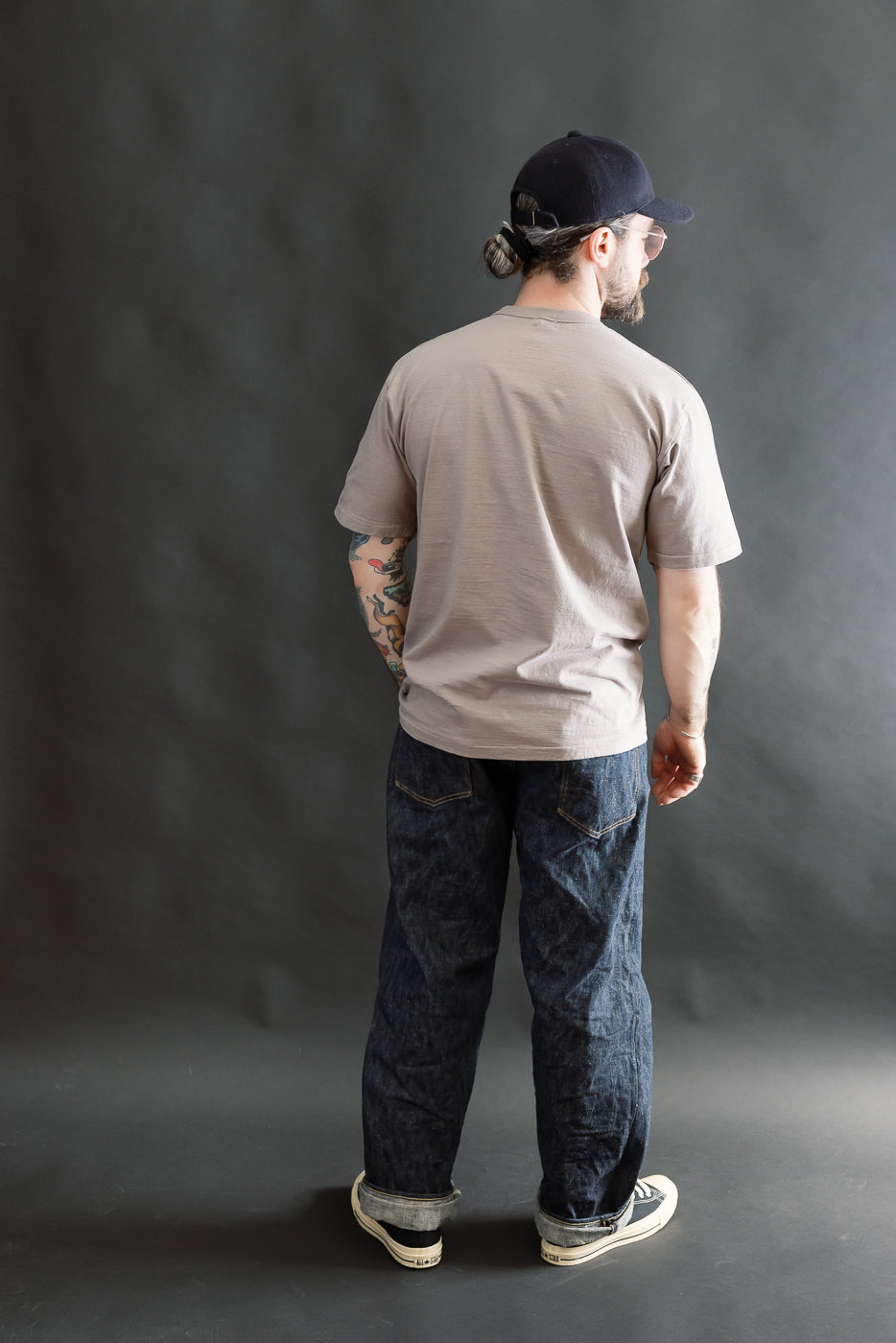 A man models the 7.7oz - Fallen Cotton Midweight T-Shirt S/S - Grey, showcasing its short sleeves and recycled cotton texture, against a gray backdrop, paired with dark jeans and sneakers.