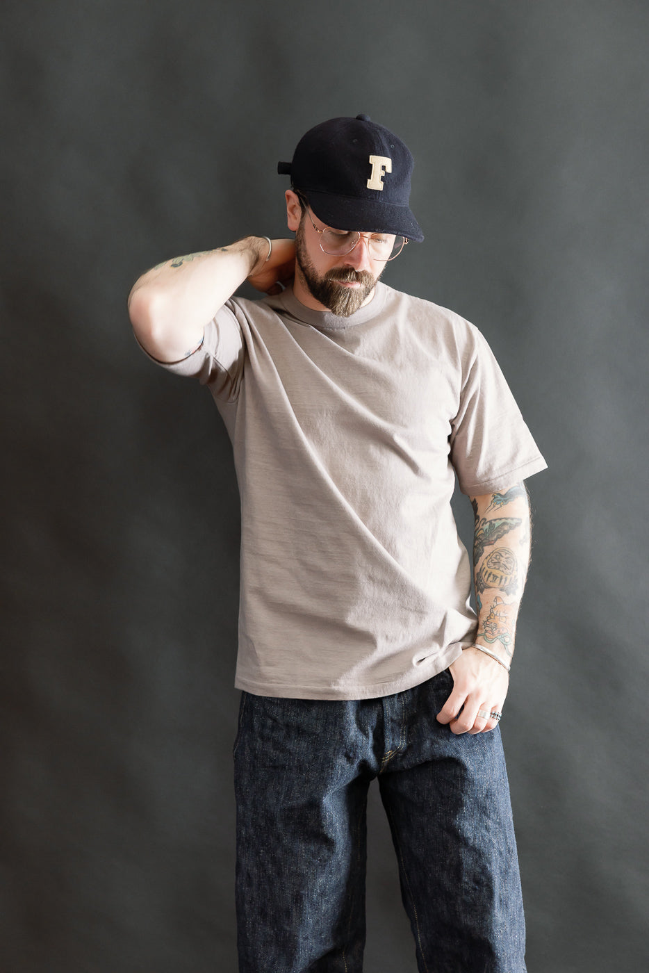 A man models the 7.7oz Fallen Cotton Midweight T-Shirt S/S - Grey, showcasing its relaxed fit, crew neck, and natural slub texture, paired with dark jeans and a black baseball cap.