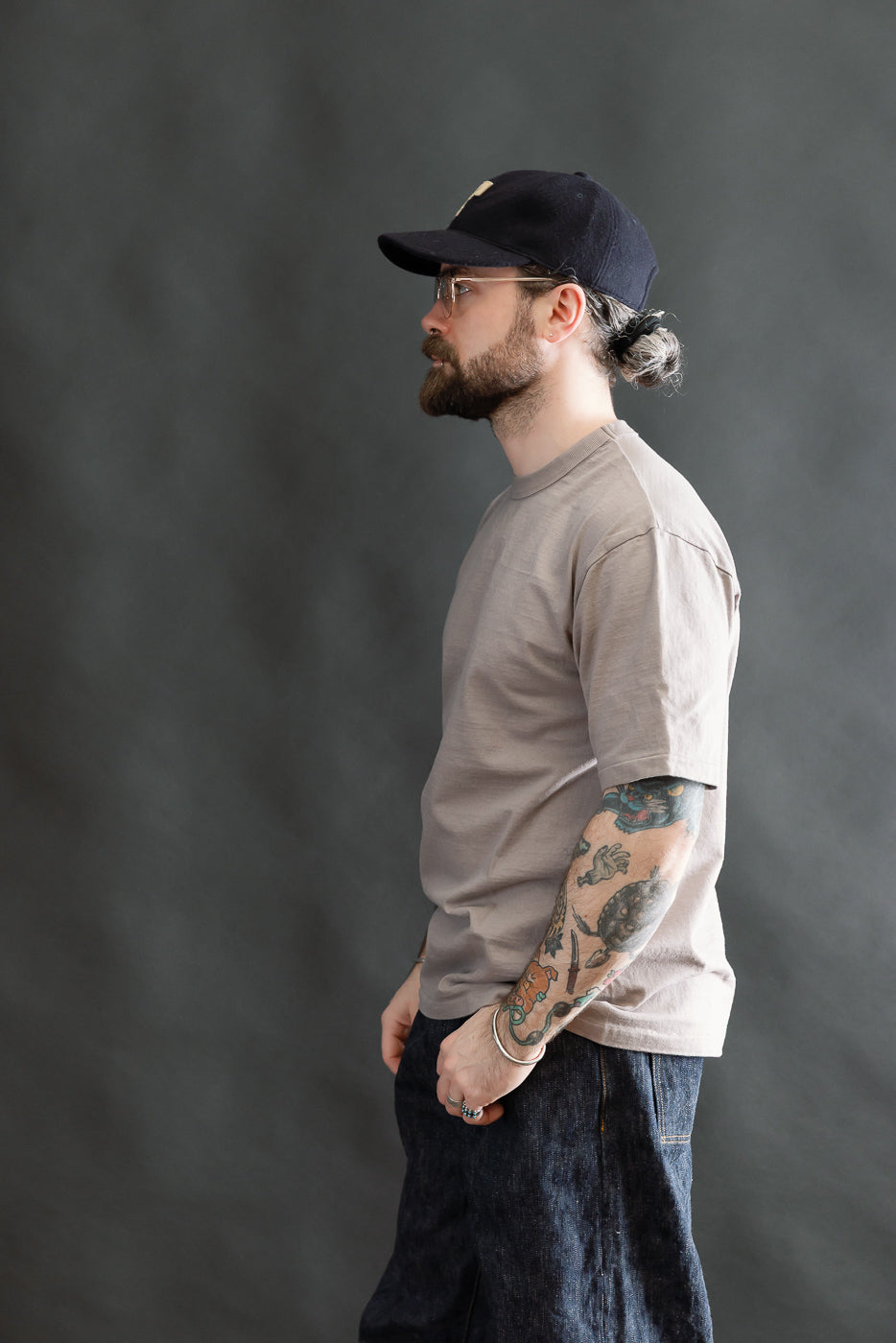 A man models the 7.7oz Fallen Cotton Midweight T-Shirt S/S - Grey, showcasing its natural uneven slub texture, round body, and classic crew-neck design, paired with dark denim jeans.