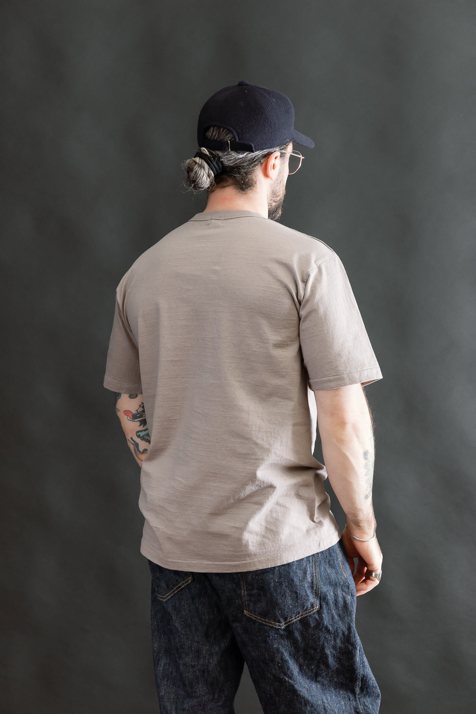 Person wearing the 7.7oz Fallen Cotton Midweight T-Shirt S/S - Grey, showcasing its short sleeves and textured recycled cotton slub jersey, paired with dark denim jeans and a black cap.