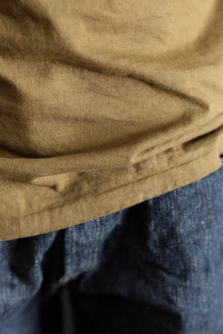 Close-up of the 7.7oz - 'Fallen Cotton' Midweight T-Shirt S/S - Khaki showcasing its textured recycled cotton slub jersey fabric, draped over blue denim jeans waistband, highlighting its casual, durable design.