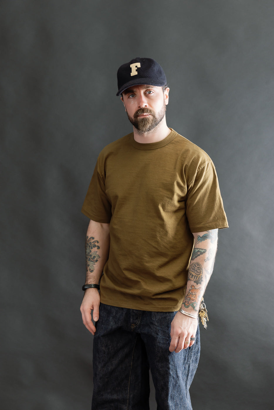 Man wearing the 7.7oz Fallen Cotton Midweight T-Shirt S/S - Khaki, showcasing its fit and texture. Styled with dark denim jeans and a navy cap, highlighting the tee's comfortable and durable design.