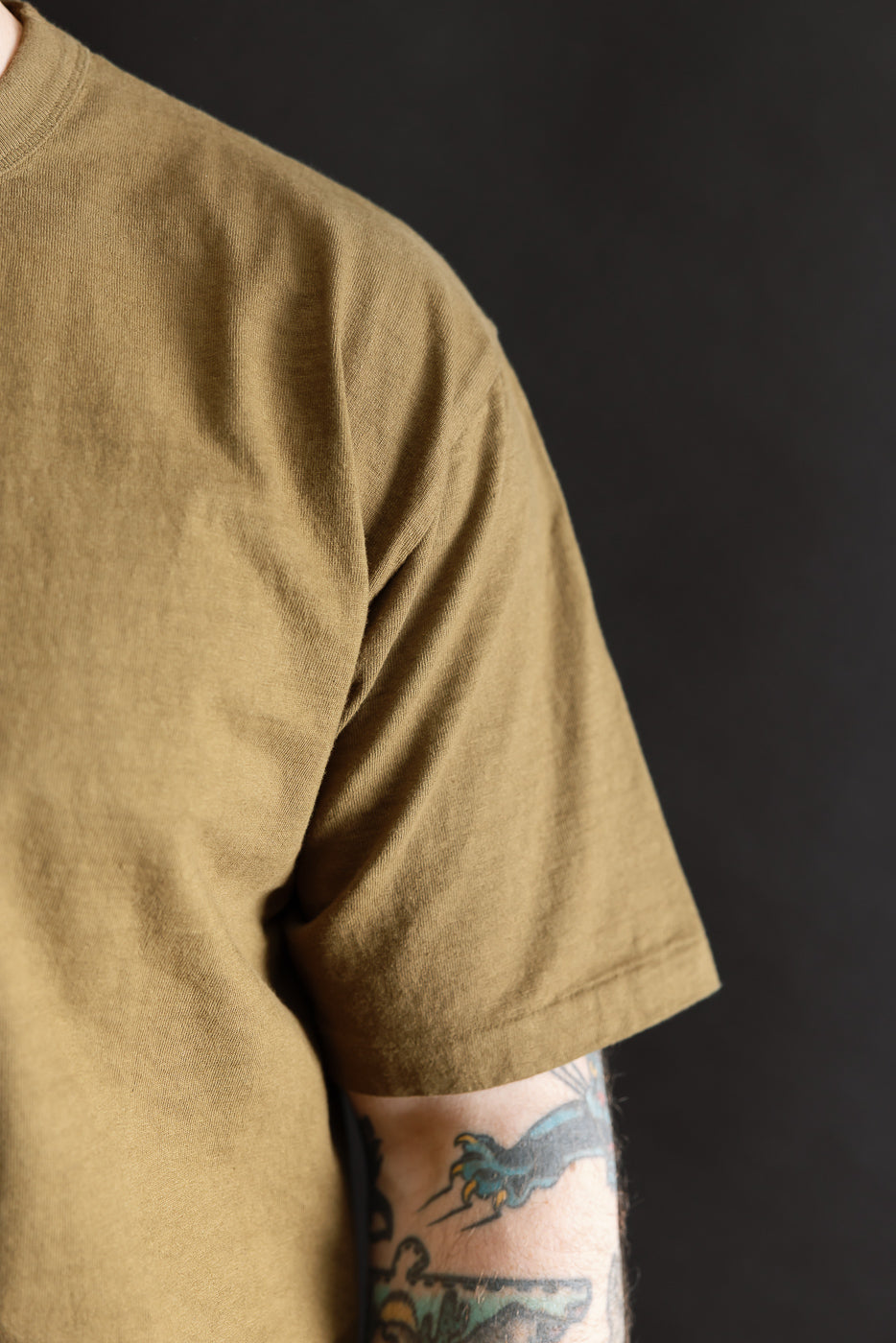 Close-up of a person wearing the Fallen Cotton Midweight T-Shirt S/S - Khaki, showcasing the textured recycled cotton slub jersey with tattooed forearm visible, emphasizing its soft, lived-in feel.