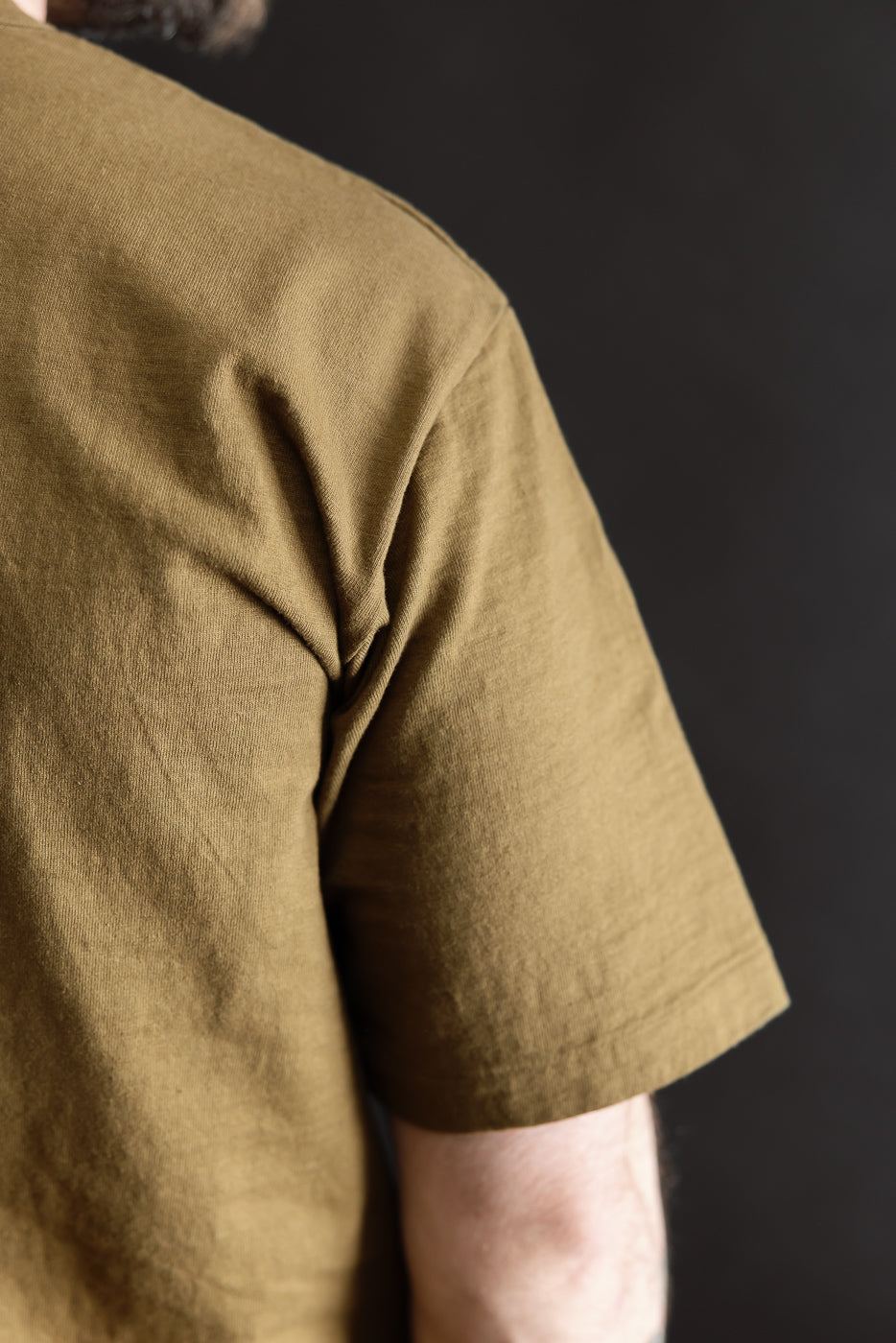 A close-up of the 7.7oz - Fallen Cotton Midweight T-Shirt S/S - Khaki showcasing its recycled cotton slub jersey texture, short sleeves, and classic binder neck detail against a dark background.