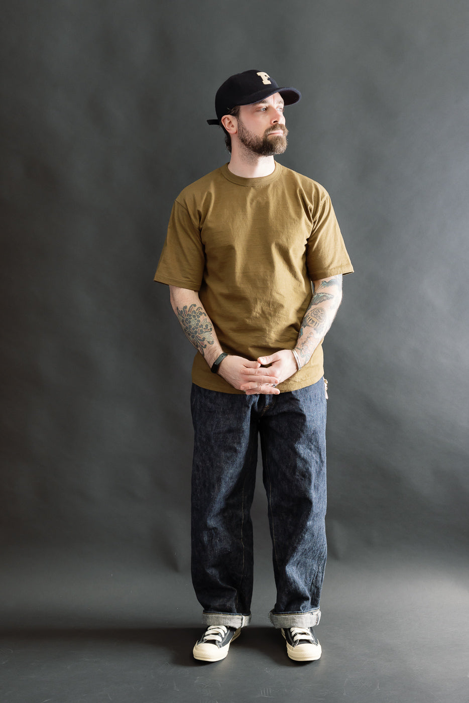 A man models the 7.7oz Fallen Cotton Midweight T-Shirt S/S - Khaki, showcasing its loose fit, rolled-up dark denim jeans, and black-and-white sneakers against a dark-gray backdrop.