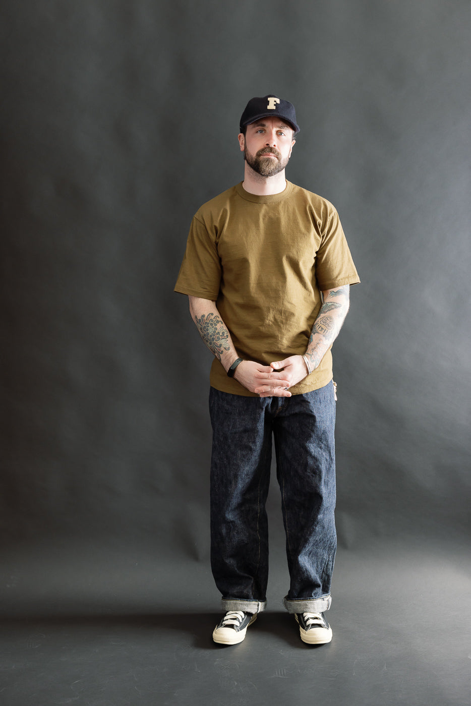 A male model wearing the 7.7oz - Fallen Cotton Midweight T-Shirt S/S - Khaki, showcasing its round body and binder neck, paired with dark cuffed jeans and black-and-white sneakers.