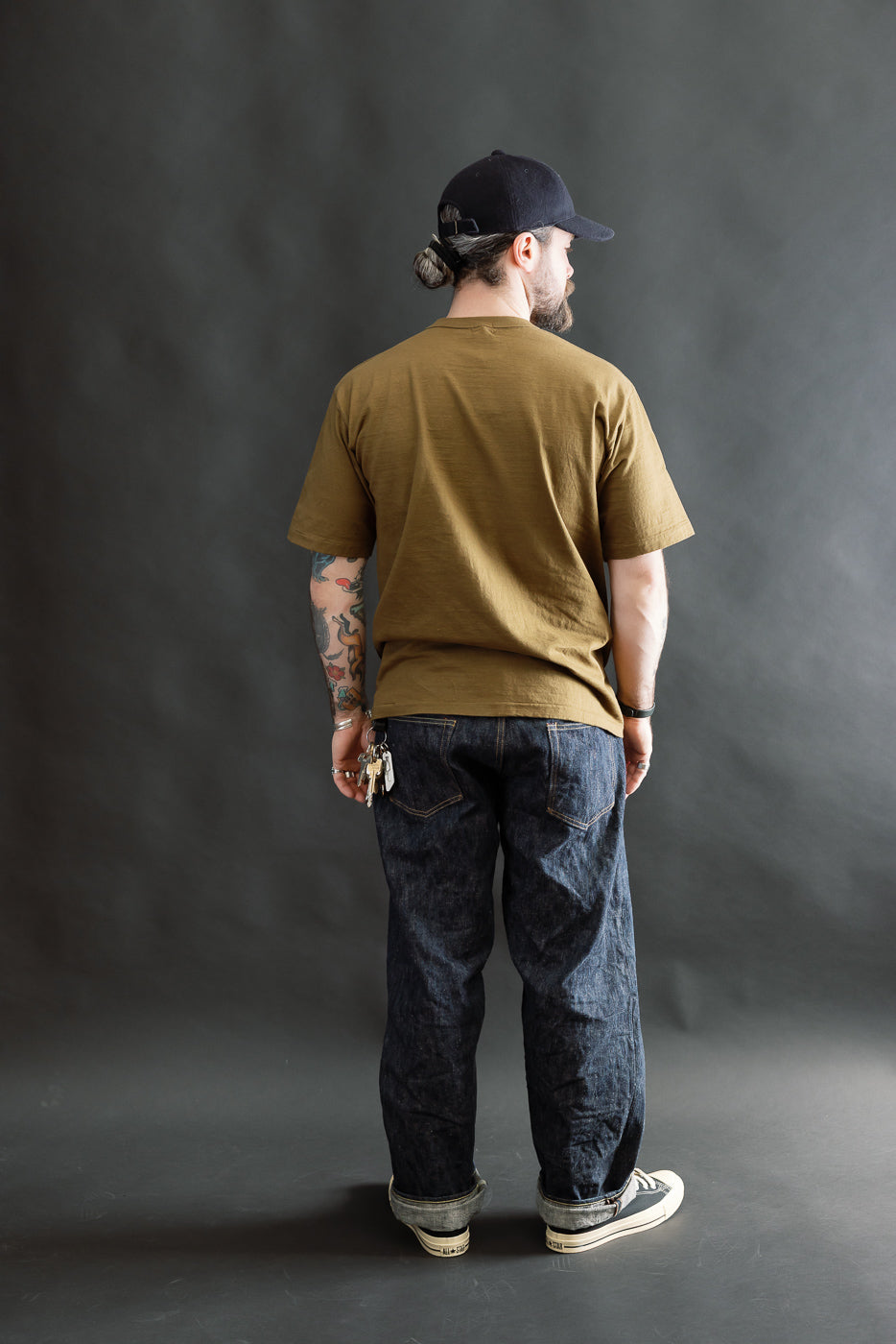 A man in jeans and sneakers showcases the 7.7oz Fallen Cotton Midweight T-Shirt S/S - Khaki, highlighting its relaxed fit and natural slub texture, with visible tattoos and a baseball cap.