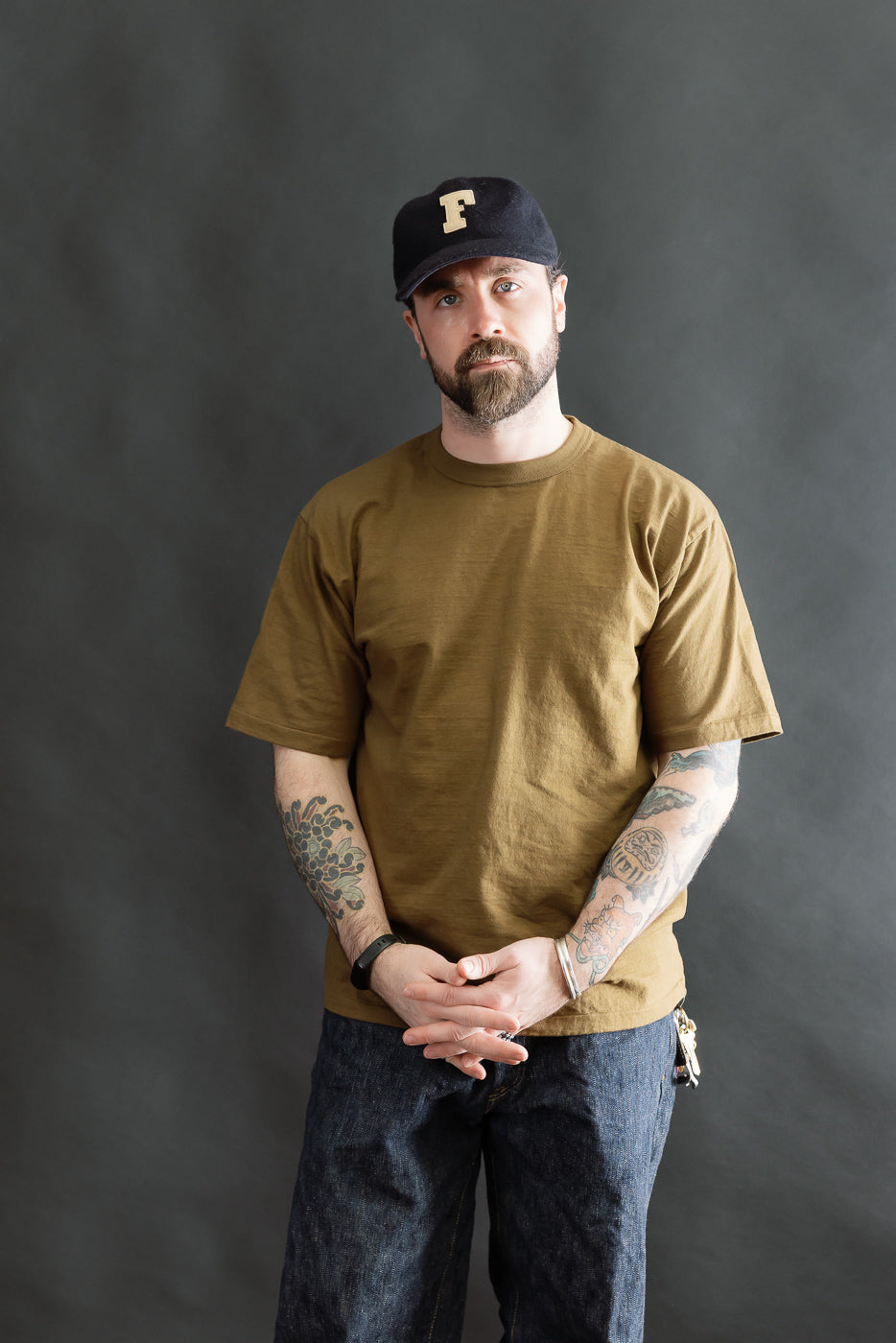 Man wearing the Fallen Cotton Midweight T-Shirt S/S - Khaki, showcasing its natural slub texture and classic fit. Styled with tattoos and jeans, embodying Dant's premium menswear ethos.