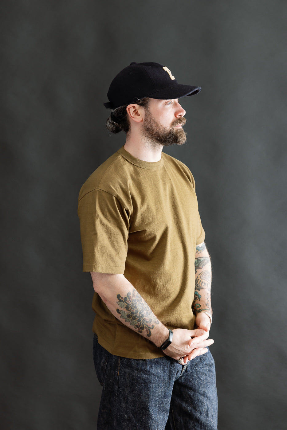 A bearded man in profile wears the 7.7oz Fallen Cotton Midweight T-Shirt S/S - Khaki, showcasing its natural slub texture and classic tee silhouette, with tattoos visible on both arms.