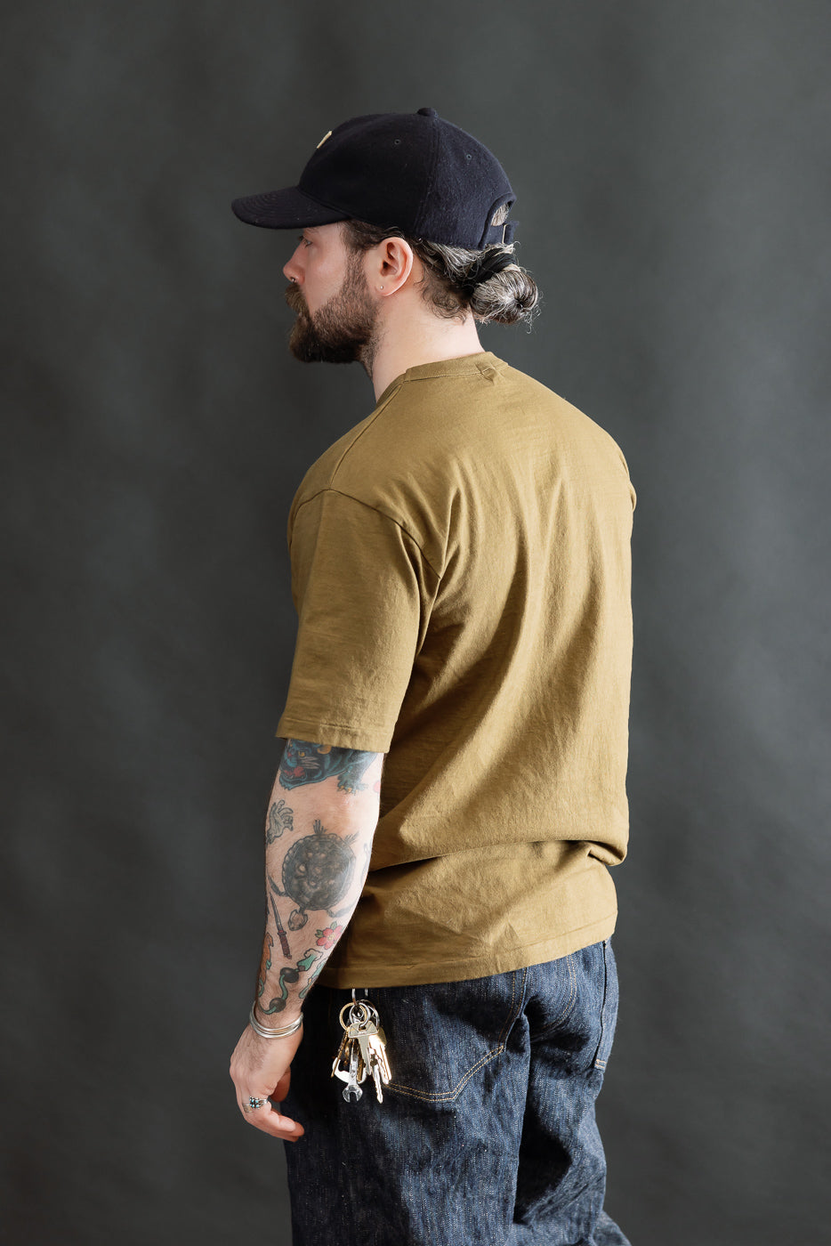 Man in profile wearing a 7.7oz - Fallen Cotton Midweight T-Shirt S/S - Khaki, showcasing the shirt's fit and texture, complemented by dark denim and tattoos, embodying Dant's high-end fashion ethos.