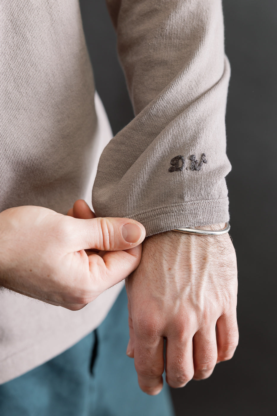 Close-up of male model's left arm wearing grey heavyweight long sleeve tee, showing sleeve cuff and embroidered logo detail. Model is 5'9