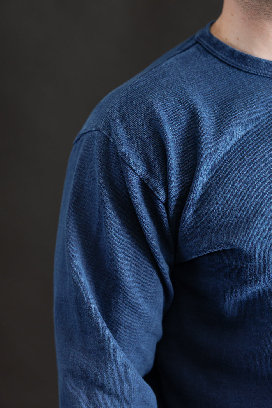 Close-up of male model's right shoulder in indigo-dyed heavyweight long sleeve tee, showing fabric texture and seam detail. Model is 5'9