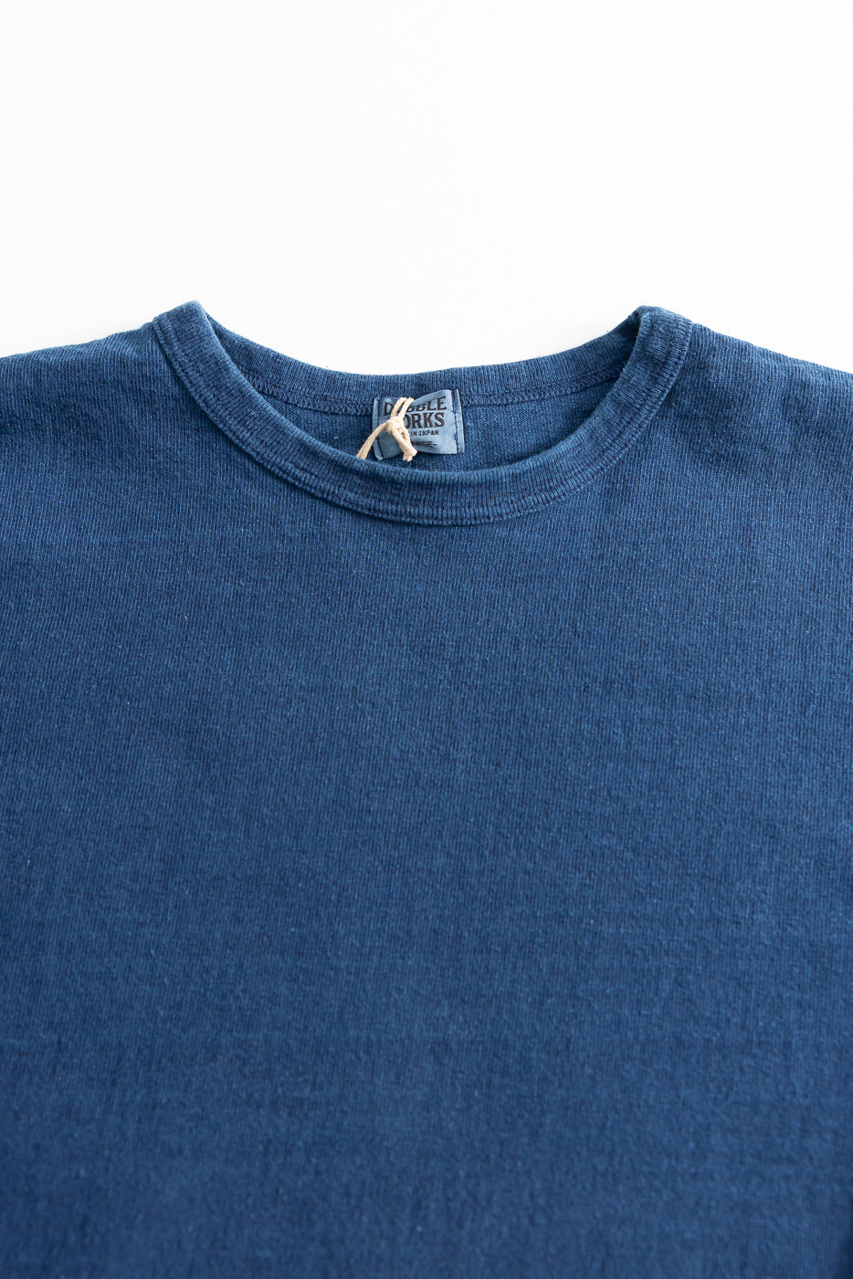 Close-up of front upper chest and collar of indigo dyed heavyweight long sleeve tee, showing ribbed crew neck, fabric texture, and visible tag detail.