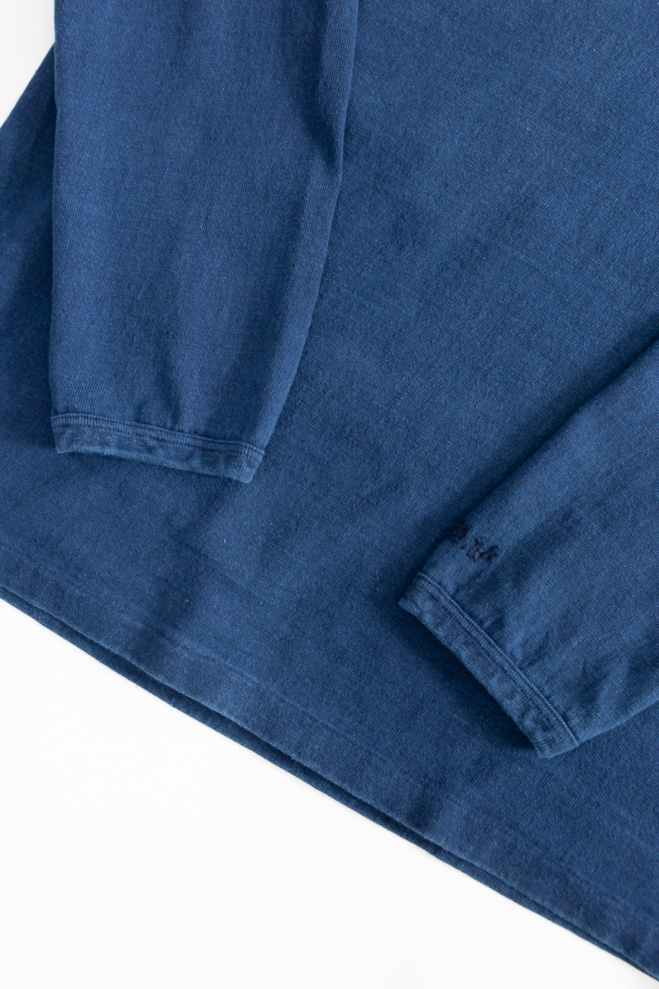 Close-up of indigo dyed heavyweight long sleeve tee, flat lay showing front hem and sleeves, fabric texture visible.