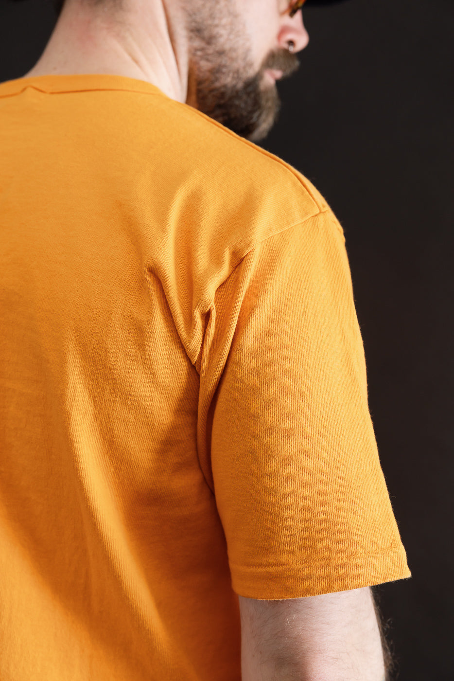 Close-up of male model's upper back and right sleeve wearing heavyweight fade orange tee, showing fabric texture and back side detail. Model is 5'9
