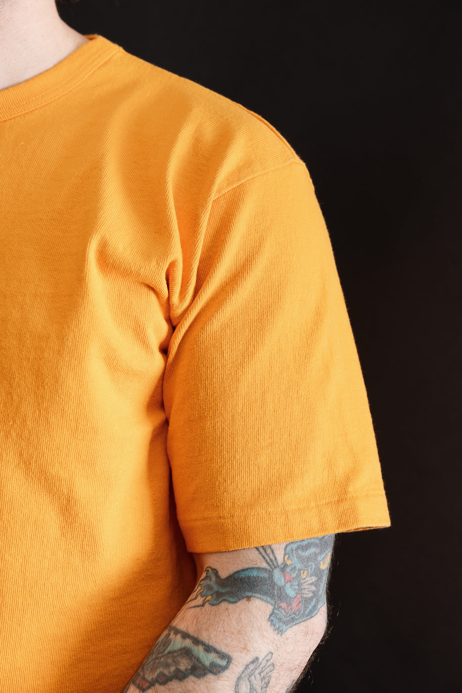 Close-up of male model’s right arm and chest in fade orange heavyweight tee, front side, showing sleeve hem and fabric texture, model is 5'9