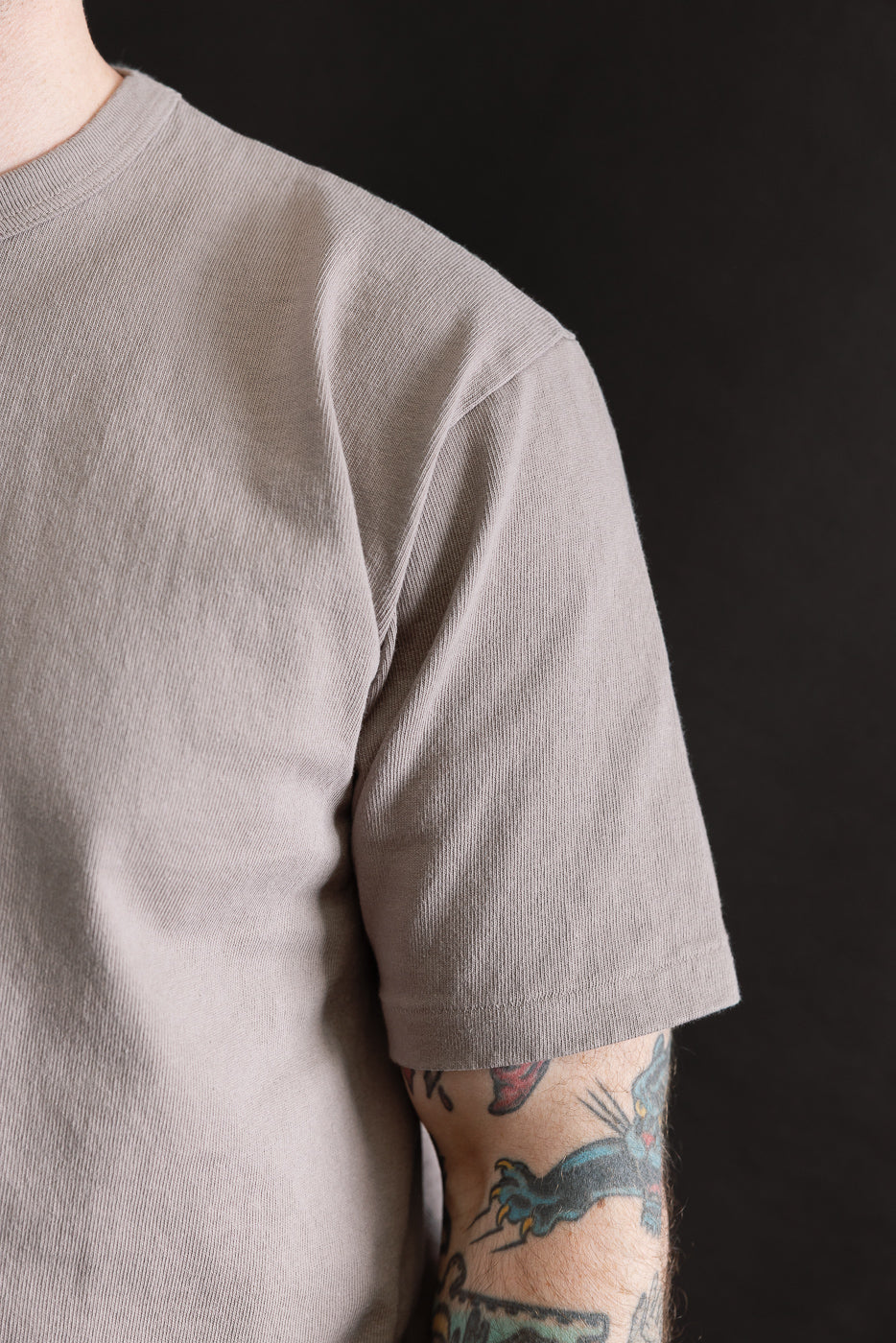 Close-up of male model's right side wearing grey heavyweight tee, showing shoulder, sleeve hem, and fabric texture. Model is 5'9