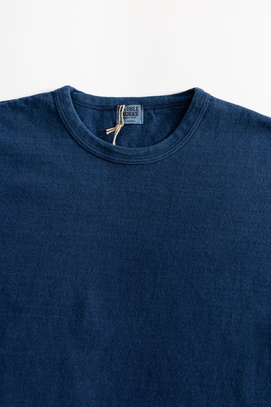Close-up of front upper chest and neckline of indigo-dyed heavyweight tee, showing ribbed crew neck and fabric texture with visible tag.