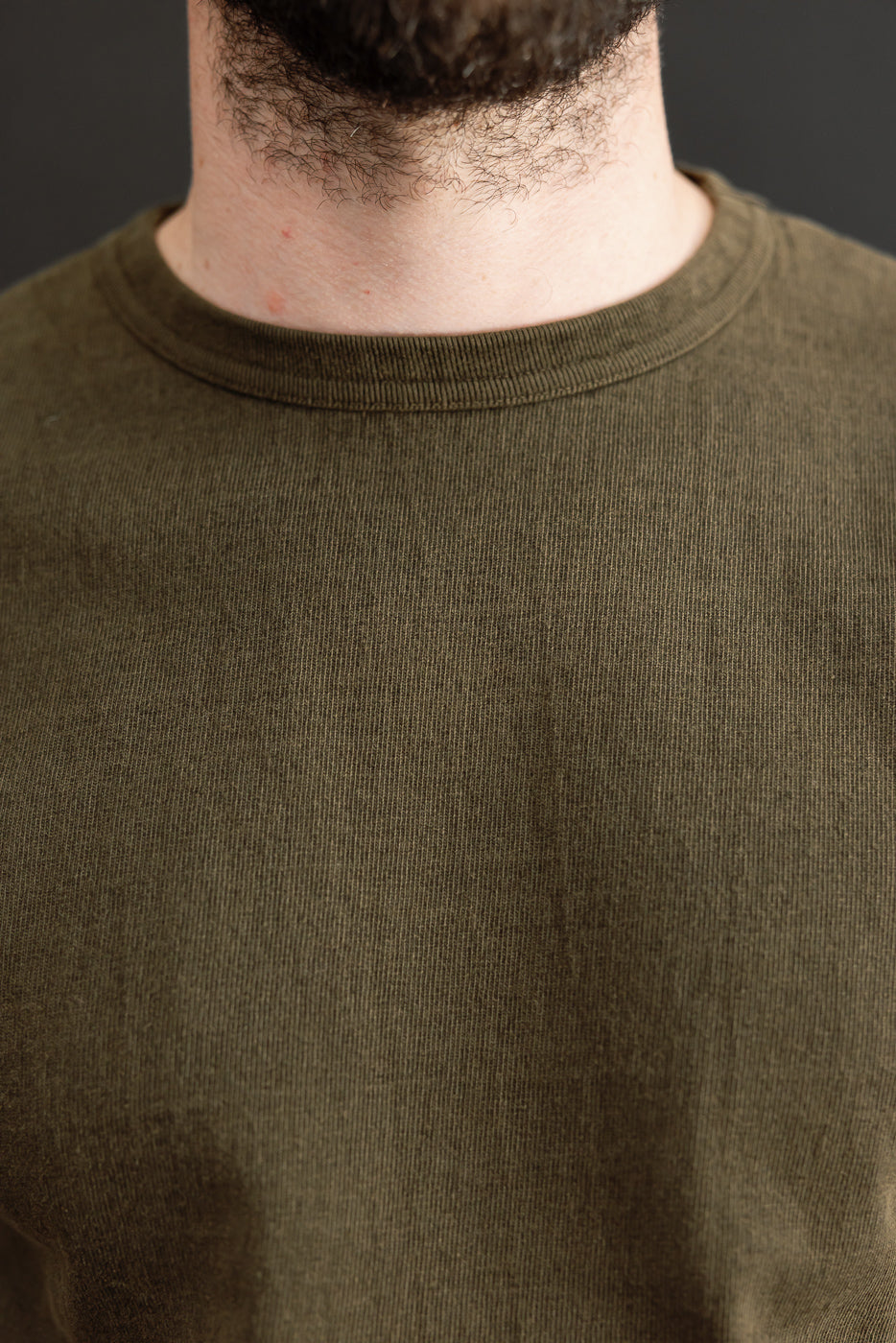 Close-up front view of olive crew neck tee on male model, showing ribbed collar and fine knit texture.