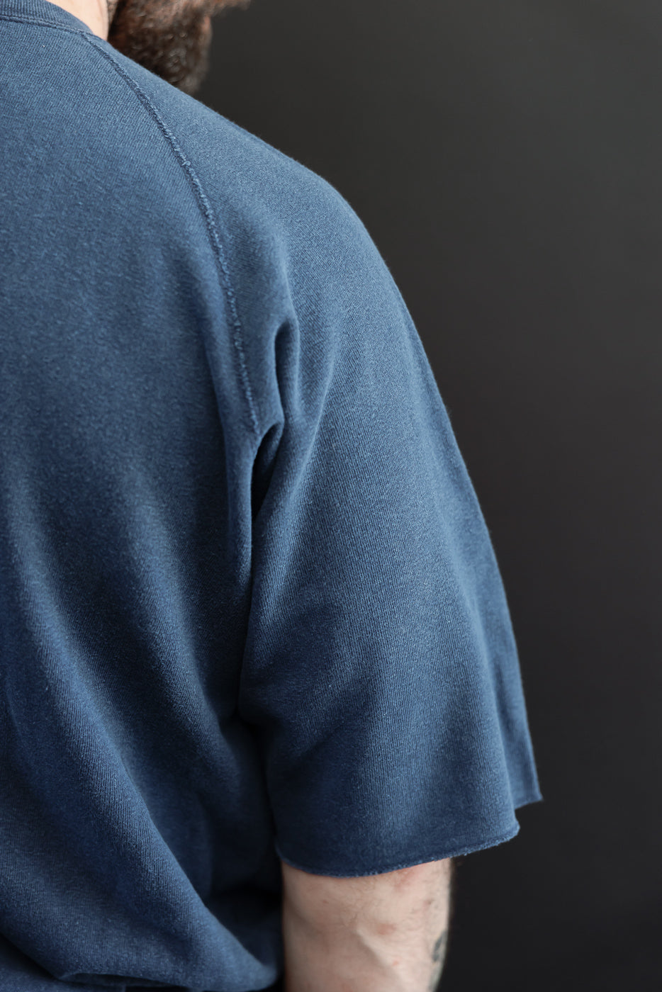 Close-up back view of male model wearing indigo short-sleeve tee, showcasing shoulder seam and fabric texture