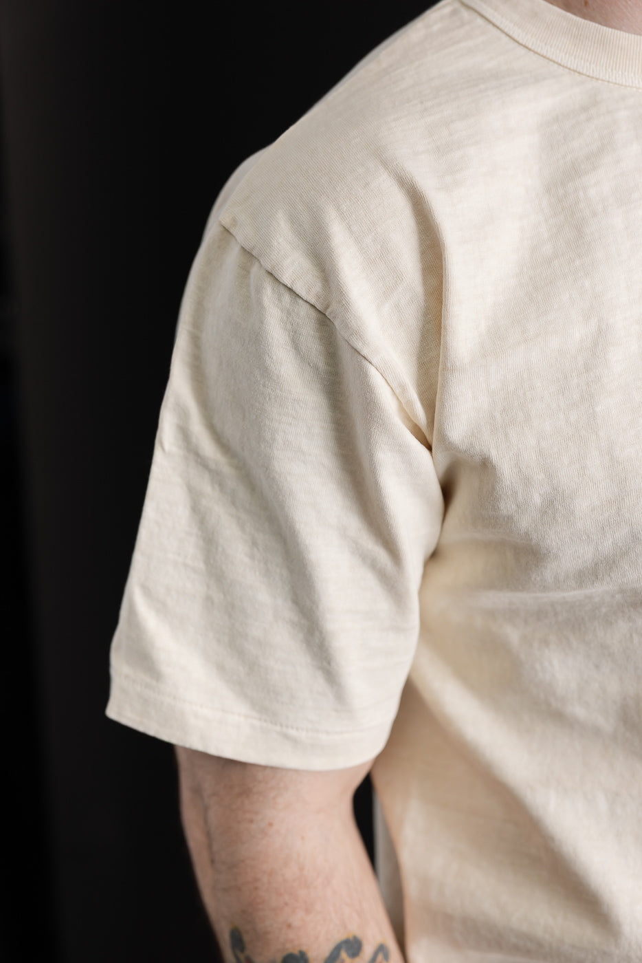 Close-up of Dubble Works Fallen Cotton Midweight Tee sleeve, cream, on male model's right side, showing fabric texture and short sleeve detail.