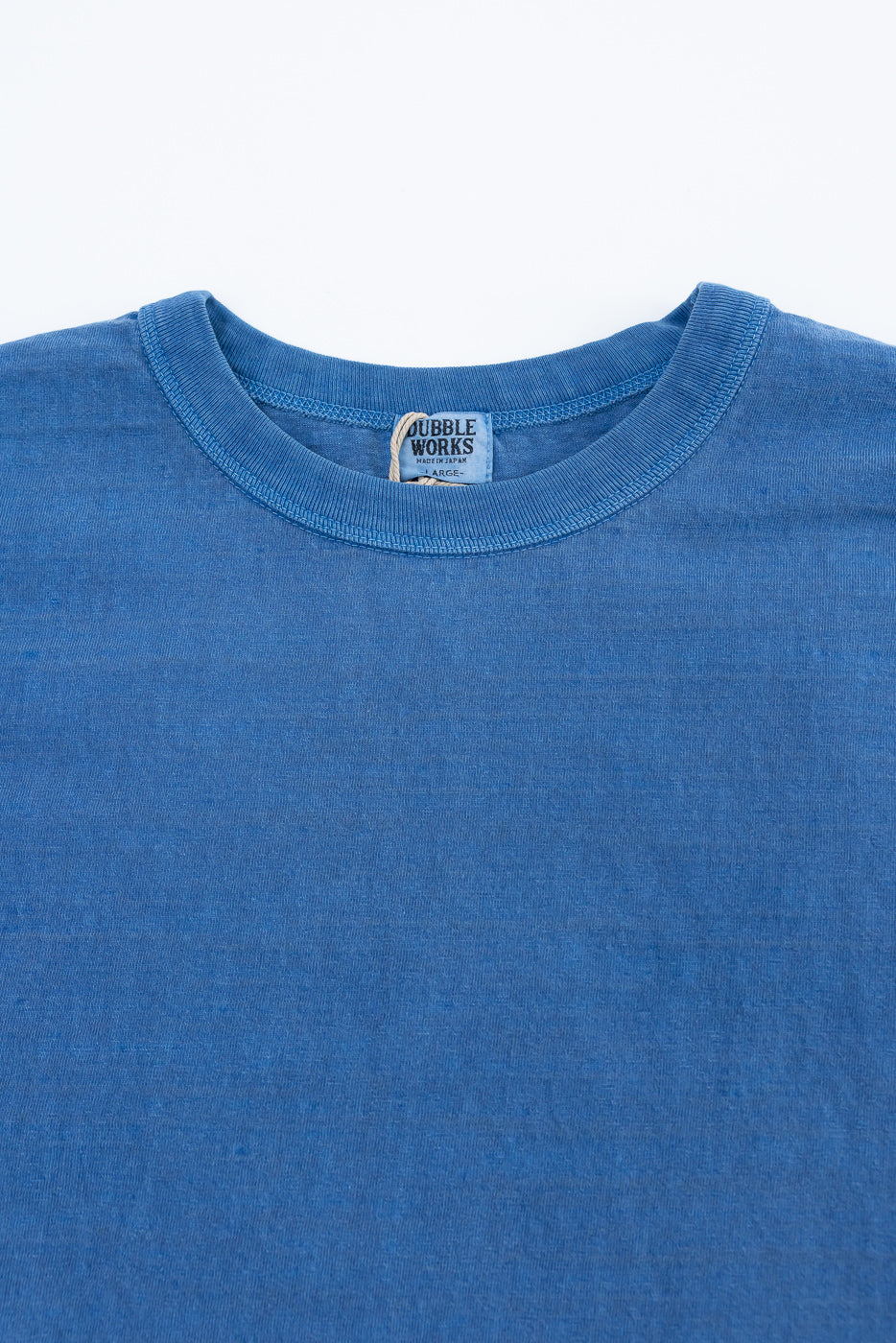 Close-up front view of Dubble Works light indigo cotton tee, showing fabric texture, ribbed crew neck, and logo tag. Contemporary casual apparel at Dant.