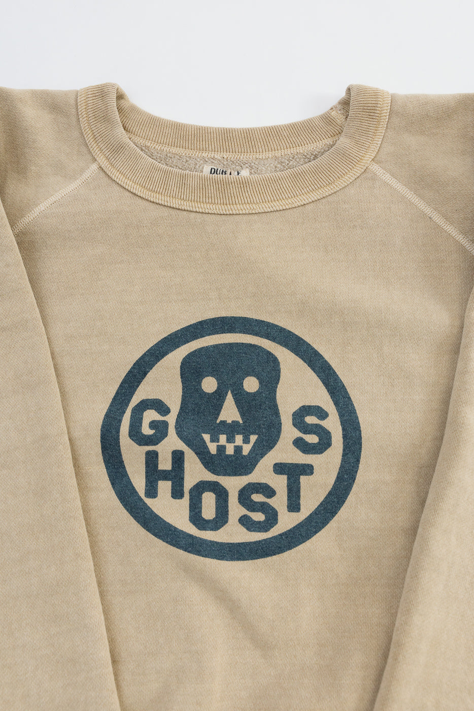Close-up of front chest graphic on beige crewneck sweatshirt, showing faded skull and 'Ghosts' print, ribbed collar, and fabric texture.