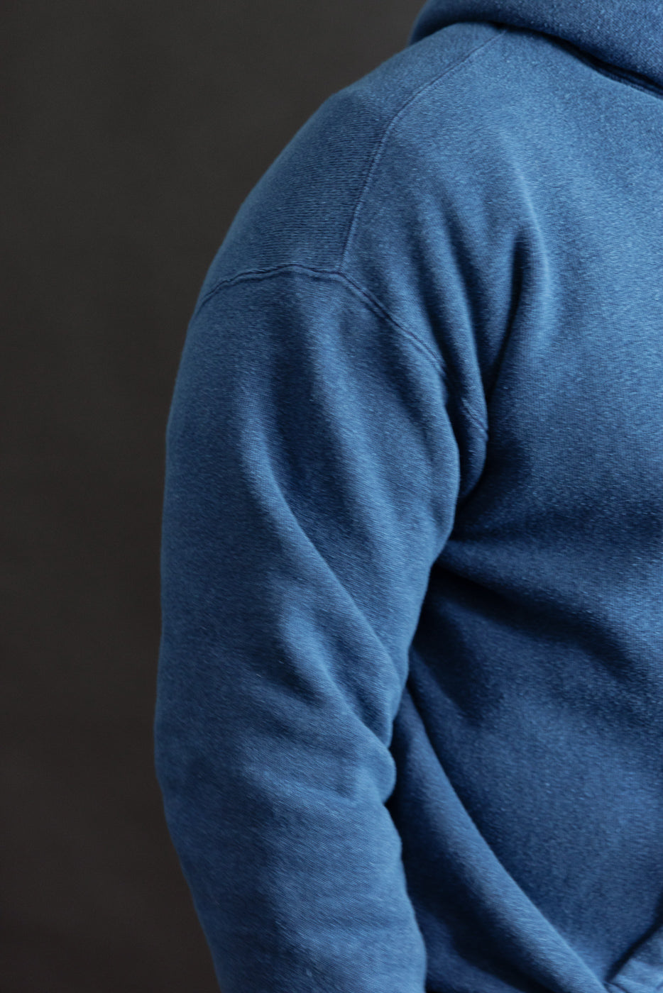 Close-up of indigo-dyed loopwheel hoodie on male model, showing left shoulder and sleeve fabric texture.