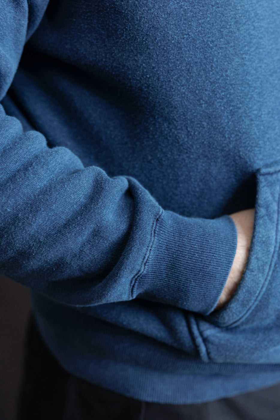 Close-up of male model's hand in kangaroo pocket, indigo-dyed sweatshirt, side view, cuff and fabric texture visible.