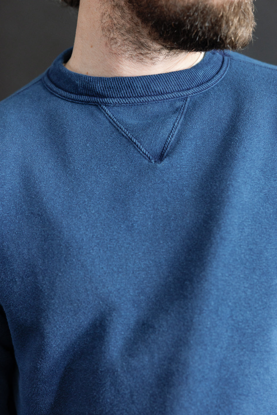 Close-up front view of indigo-dyed loopwheel sweatshirt collar on male model, highlighting fabric texture and V-stitch detail. Model is 5'9