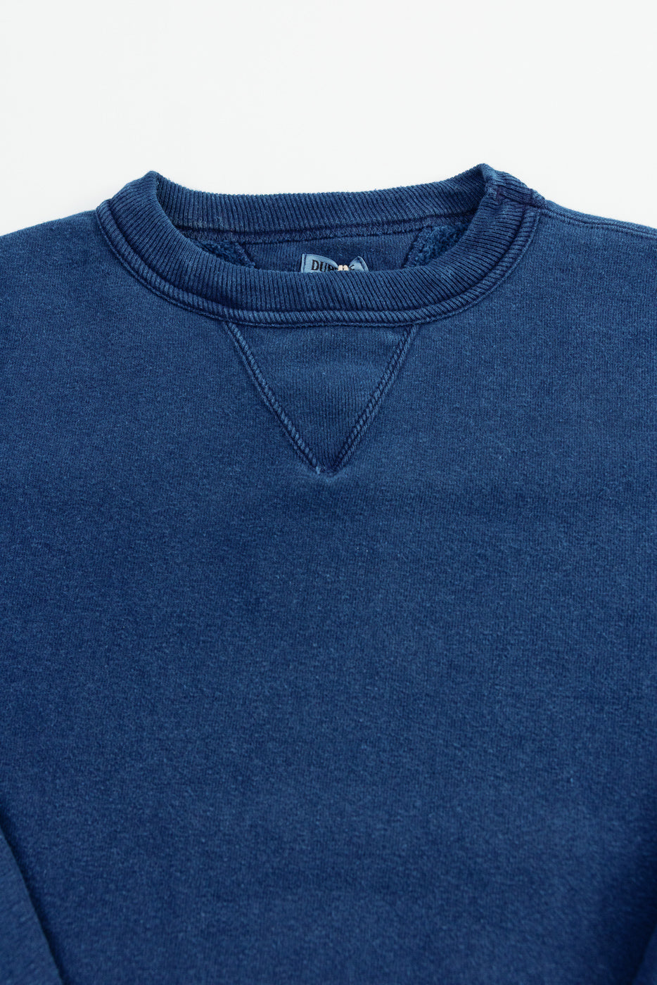 Close-up of the front upper chest and neckline of an indigo-dyed loopwheel sweatshirt, highlighting ribbed crewneck collar, V-stitch detail, and fabric texture.