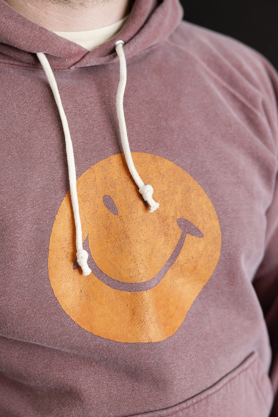 Close-up of front chest on faded bordeaux hoodie with yellow smiley face print, showing drawstrings. Male model, 5'9