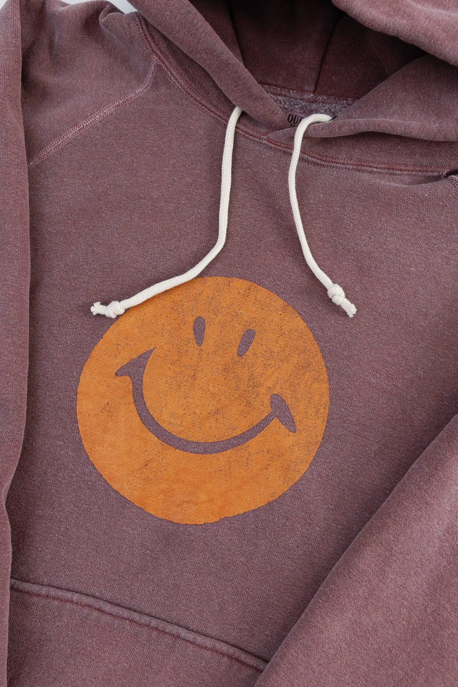 Close-up of front chest on bordeaux faded print hoodie with orange smiley face graphic, drawstrings, and visible fabric texture.