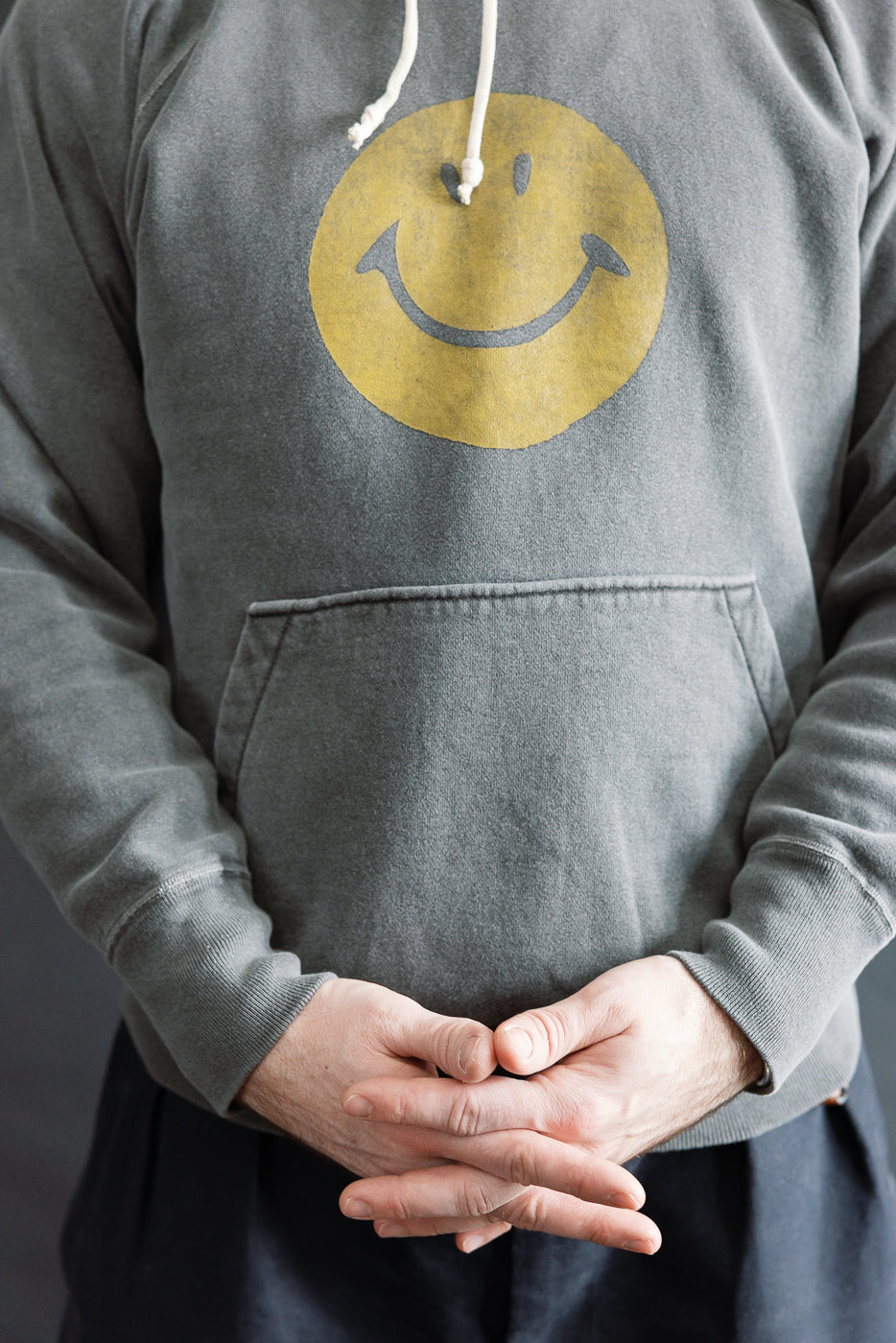 Close-up front view of male model 5'9, 165lbs, athletic build in grey faded print hoodie with yellow smiley graphic and kangaroo pocket.