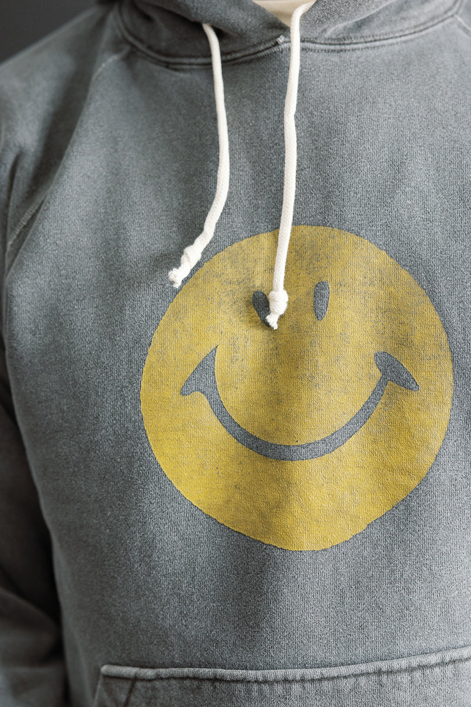Close-up front of men's faded grey hoodie with yellow smiley face print, showing chest and kangaroo pocket details. Model is male, 5'9