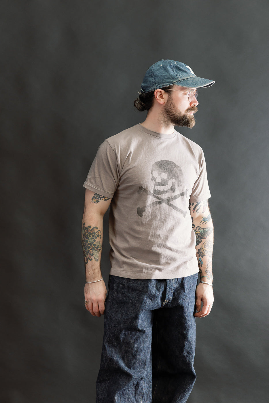 Model wearing the 5.4oz Raffy Tenjiku Printed T-Shirt Skull & Crossbone - Grey, showcasing colorful tattooed forearms, clear-framed glasses, and a denim baseball cap, paired with loose-fitting dark denim jeans.