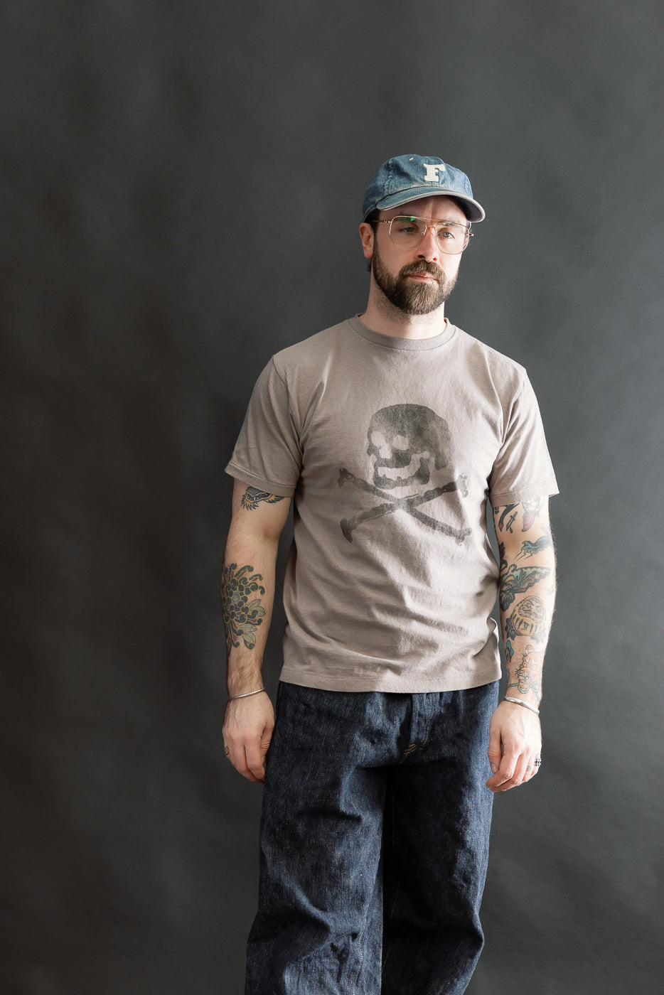 A bearded man models the 5.4oz Raffy Tenjiku Printed T-Shirt Skull & Crossbone in grey, showcasing its vintage-inspired design, tubular construction, and distinctive skull-and-crossbones motif.