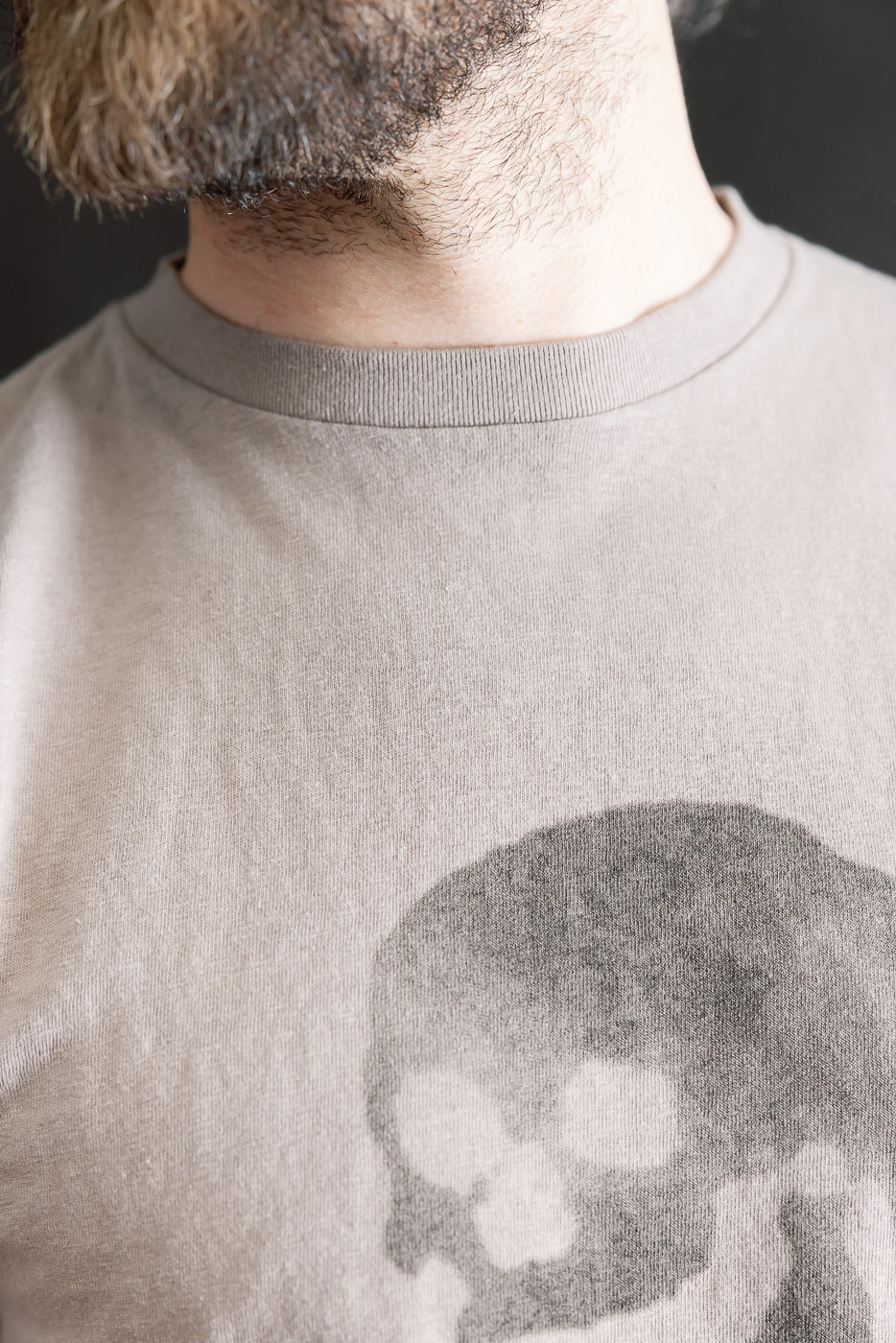 Close-up of a model wearing the 5.4oz Raffy Tenjiku Printed T-Shirt Skull & Crossbone - Grey, showcasing a stylized skull graphic on the light-colored, vintage-inspired T-shirt with a nep-like texture.