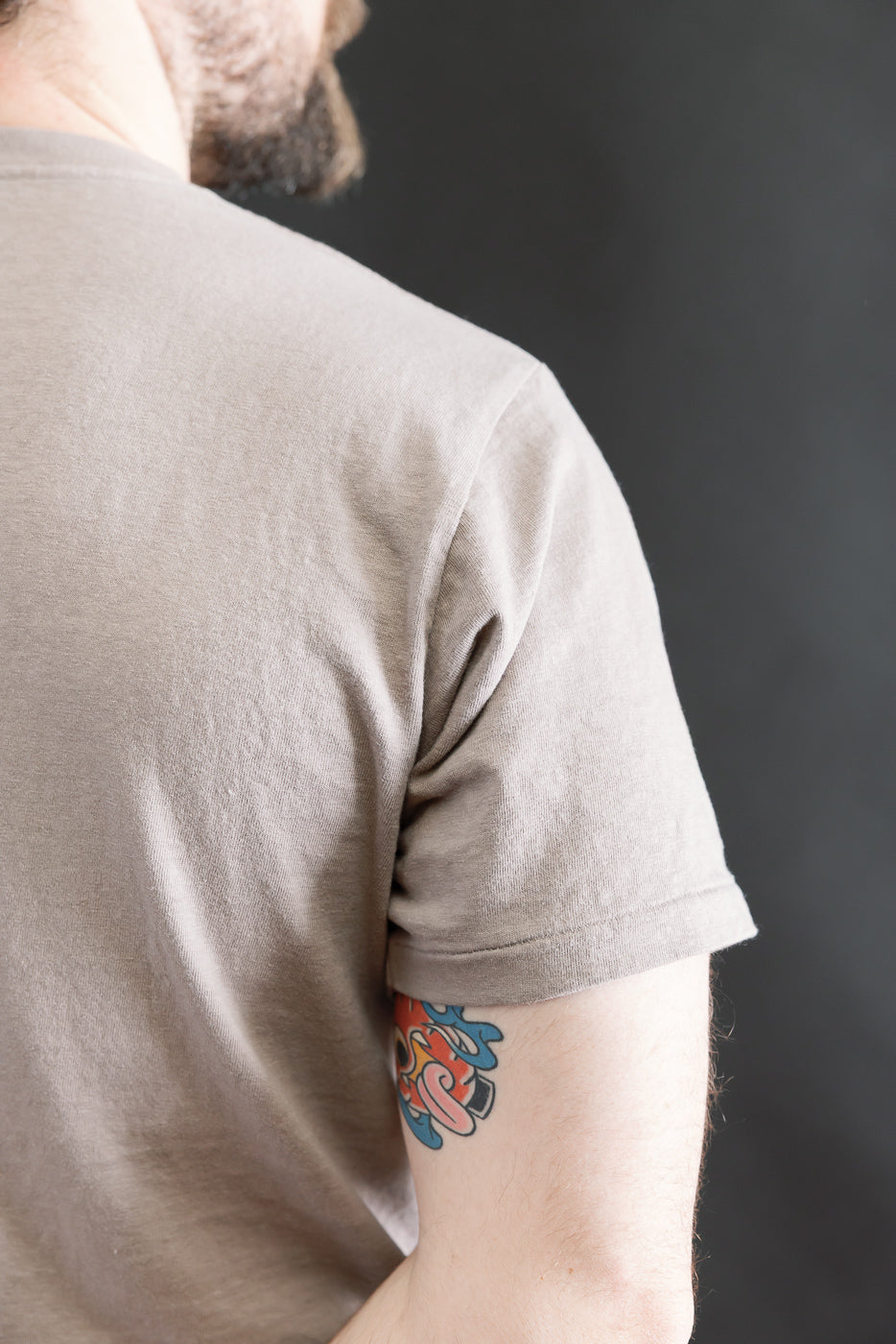 The image features the back and right upper arm of a person wearing the 5.4oz Raffy Tenjiku Printed T-Shirt Skull & Crossbone in grey, showcasing its seamless tubular knit and colorful arm tattoo.