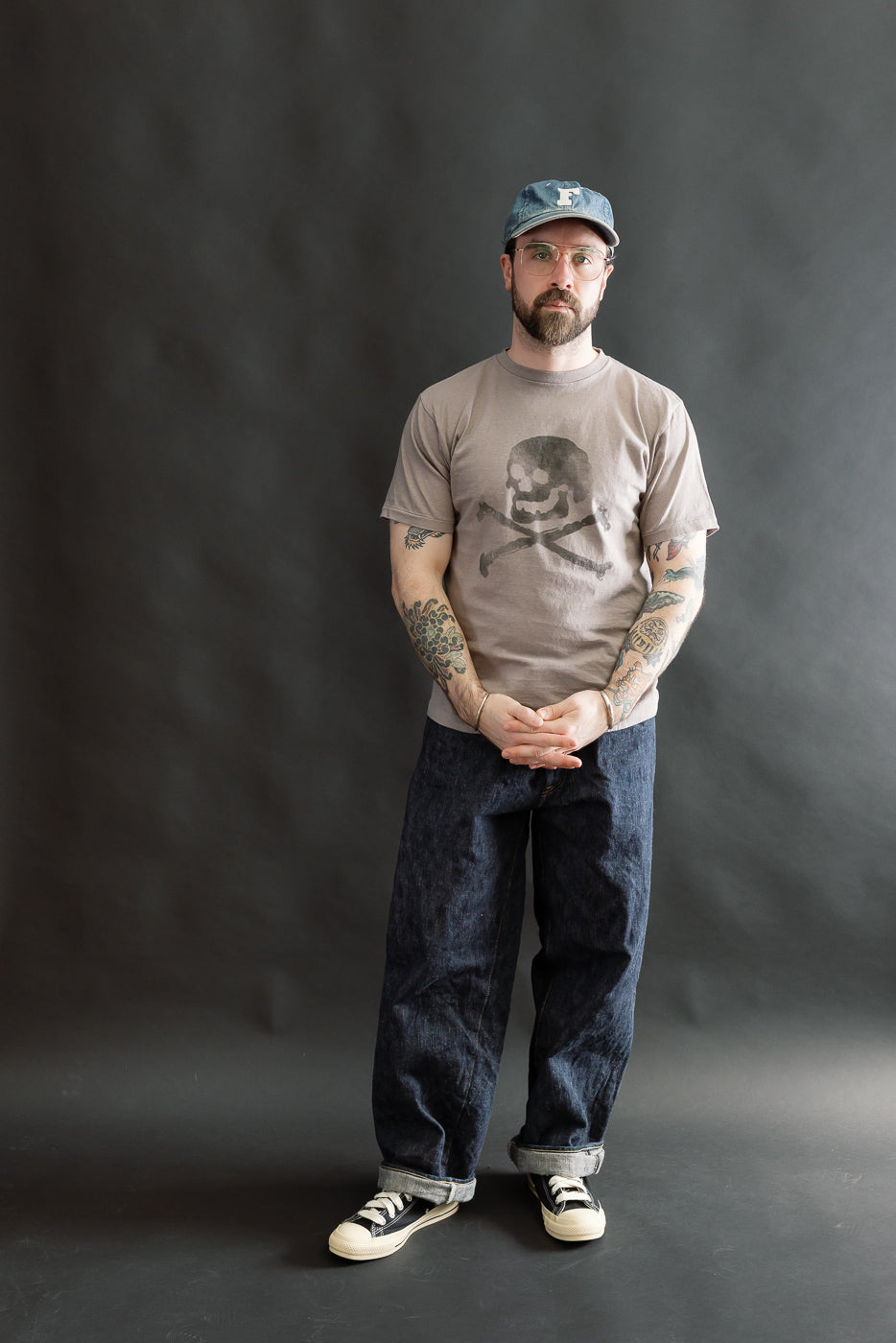 Man in a gray 5.4oz Raffy Tenjiku Printed T-Shirt Skull & Crossbone, styled with rolled-cuff denim jeans and canvas sneakers, showcasing colorful tattooed forearms and a denim cap.