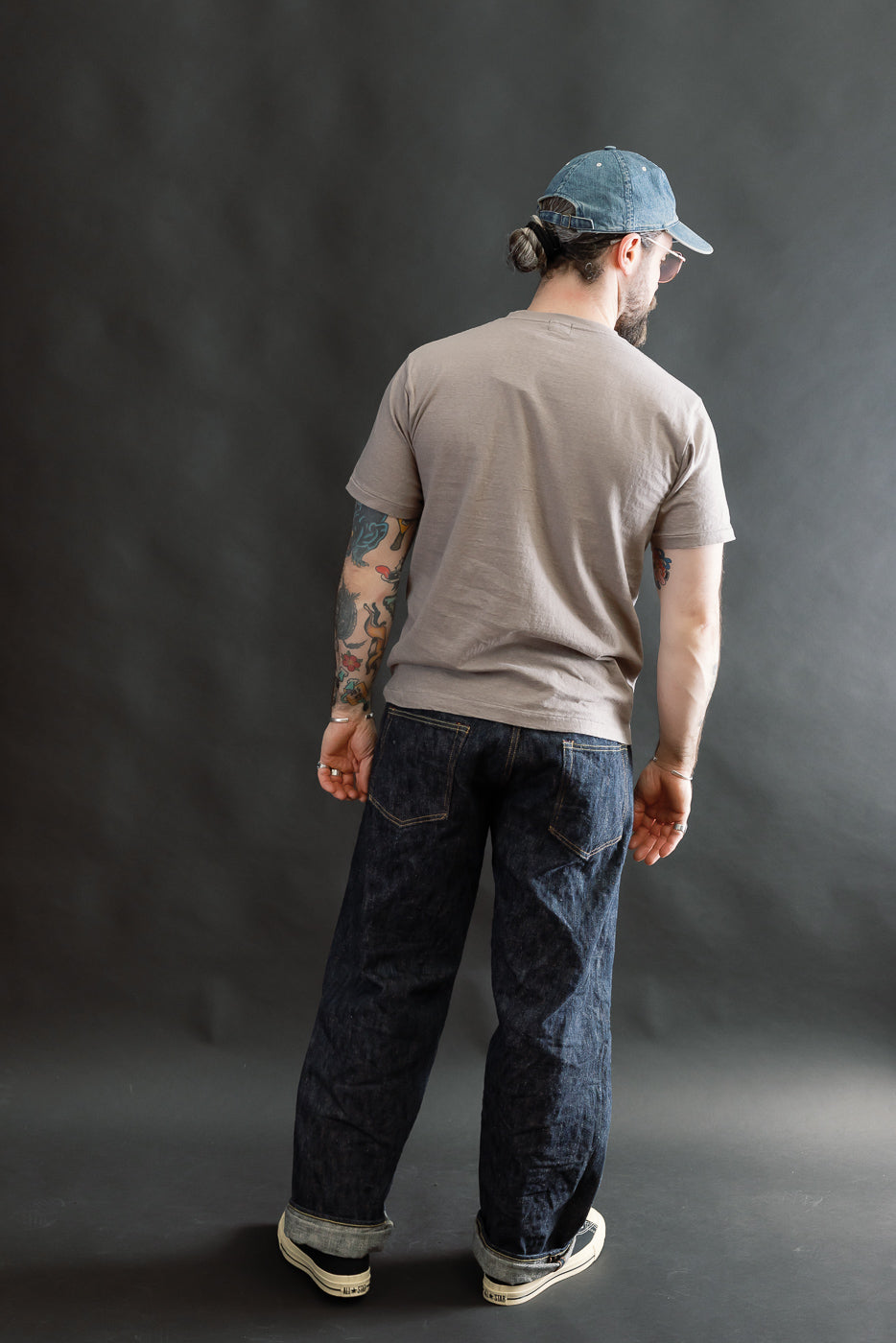 A man models the 5.4oz Raffy Tenjiku Printed T-Shirt Skull & Crossbone - Grey, showcasing its vintage-inspired design with tubular construction, paired with denim jeans and white sneakers.