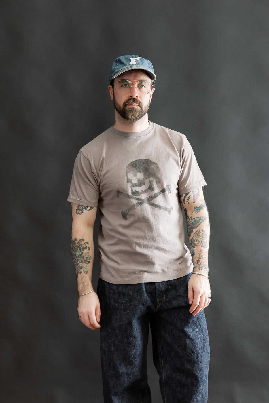 Man wearing the 5.4oz Raffy Tenjiku Printed T-Shirt Skull & Crossbone - Grey, showcasing its vintage-inspired design with tubular construction and discharge print, paired with dark indigo jeans and silver accessories.