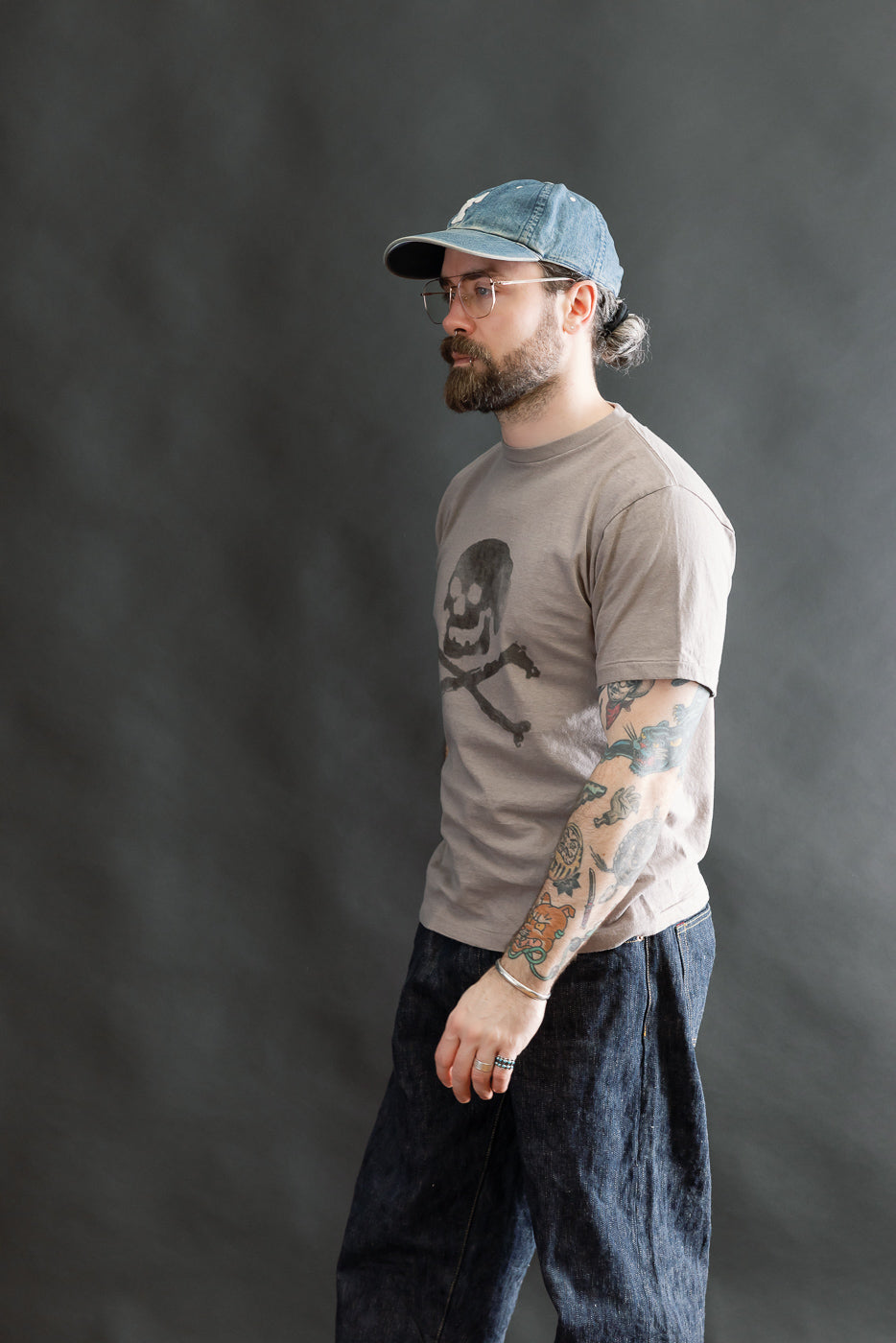 Bearded man models the 5.4oz Raffy Tenjiku Printed T-Shirt Skull & Crossbone - Grey, highlighting its vintage-inspired design with tubular knit and discharge print, styled with dark denim and accessories.
