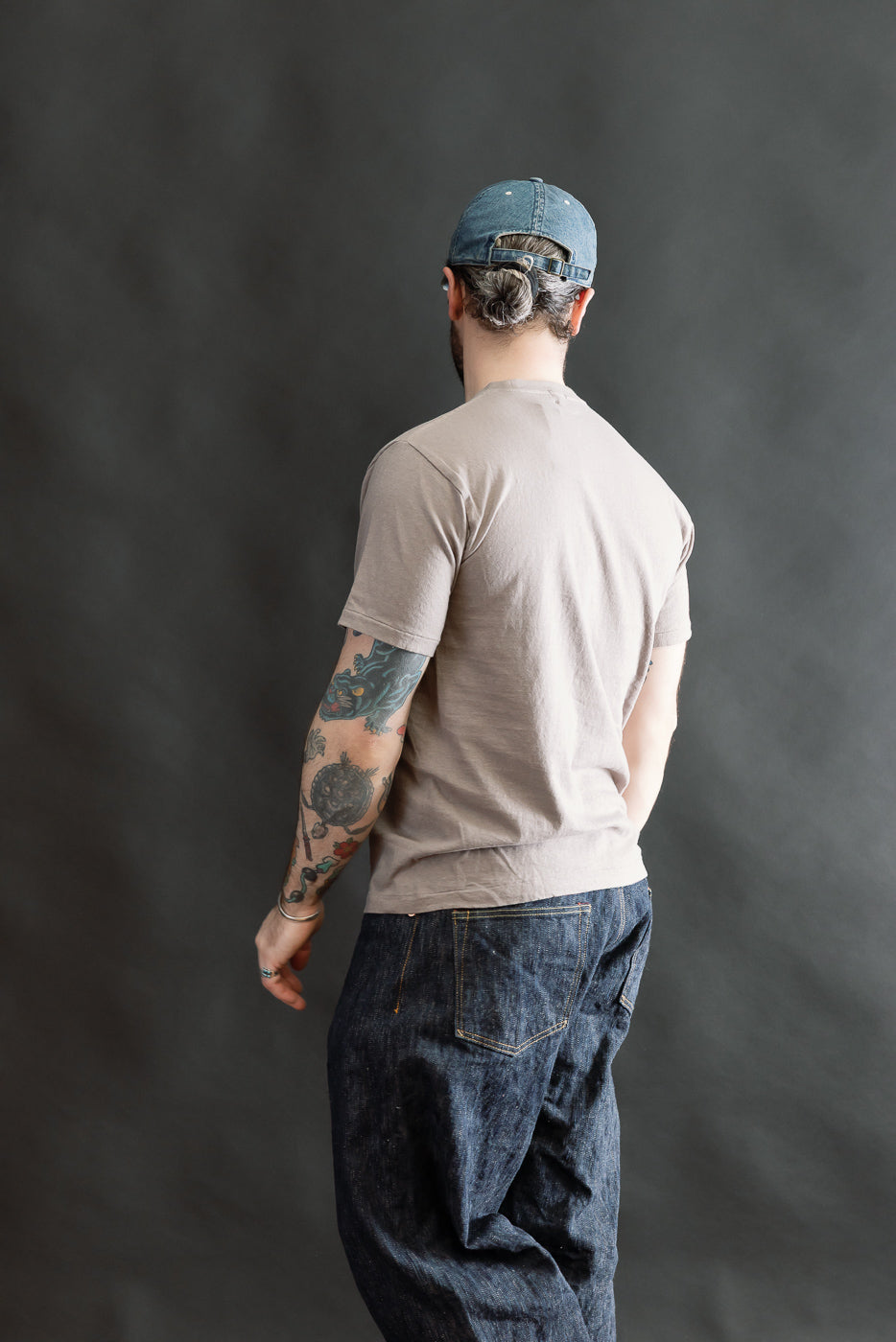 Man in light taupe 5.4oz Raffy Tenjiku Printed T-Shirt Skull & Crossbone - Grey, with dark denim jeans, blue baseball cap, showcasing tattooed arm, embodying a vintage-inspired style.