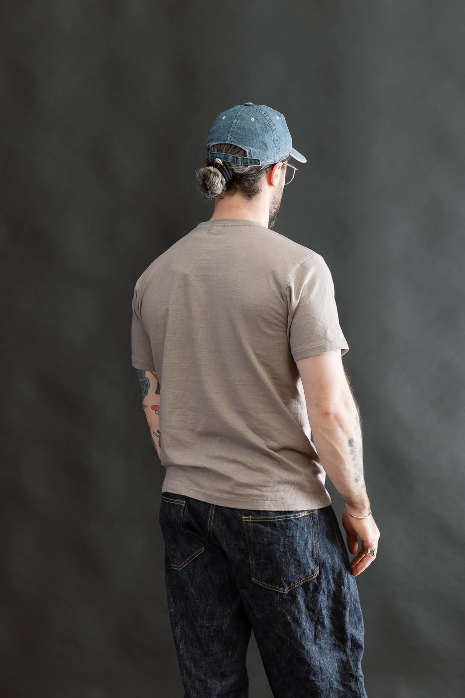 Back view of a male model wearing the 5.4oz Raffy Tenjiku Printed T-Shirt Skull & Crossbone - Grey, showcasing its seamless tubular construction and vintage-inspired fit, paired with jeans and a backward cap.