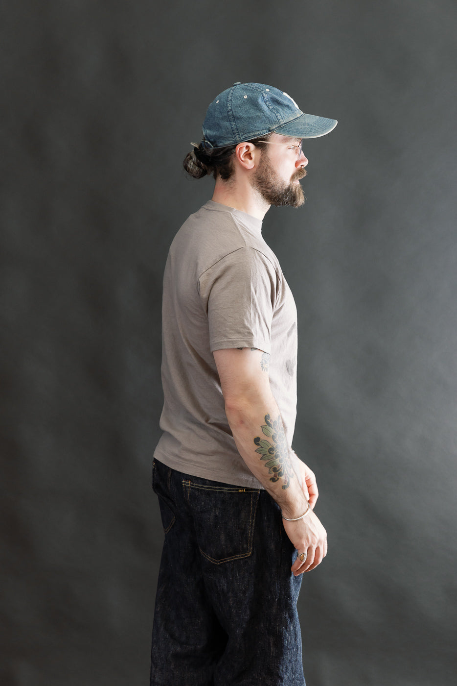 Side-profile of a man wearing the 5.4oz Raffy Tenjiku Printed T-Shirt Skull & Crossbone in grey, showcasing its vintage-inspired fit and texture, paired with dark denim jeans and a baseball cap.