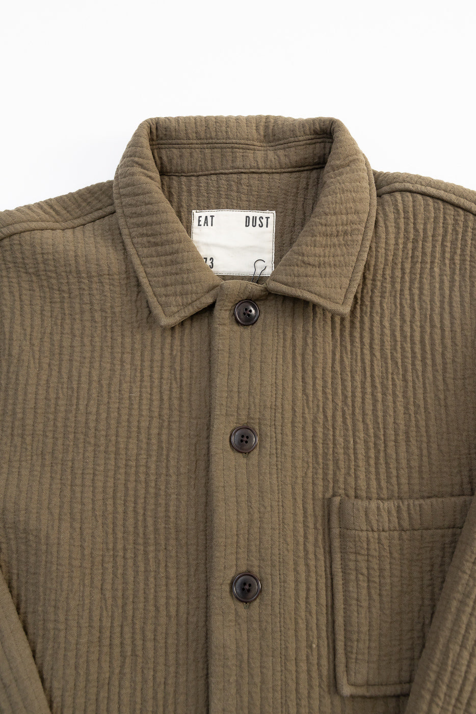 Close-up front of Eat Dust Cana shirt in fossil green, showing textured jacquard fabric, buttons, pocket, collar, and brand tag.