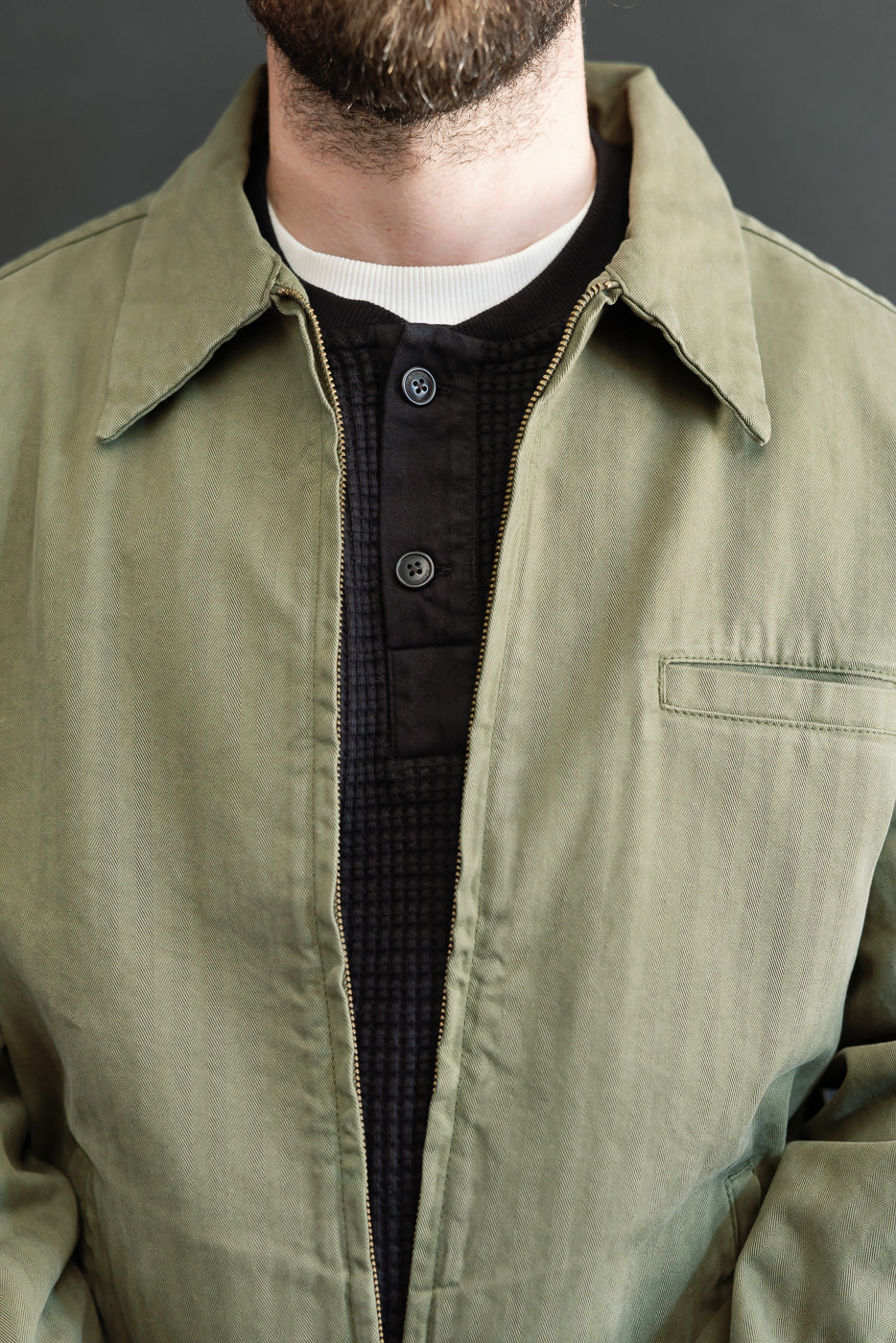 Close-up front view of an olive green canvas jacket over a black buttoned waffle knit shirt.